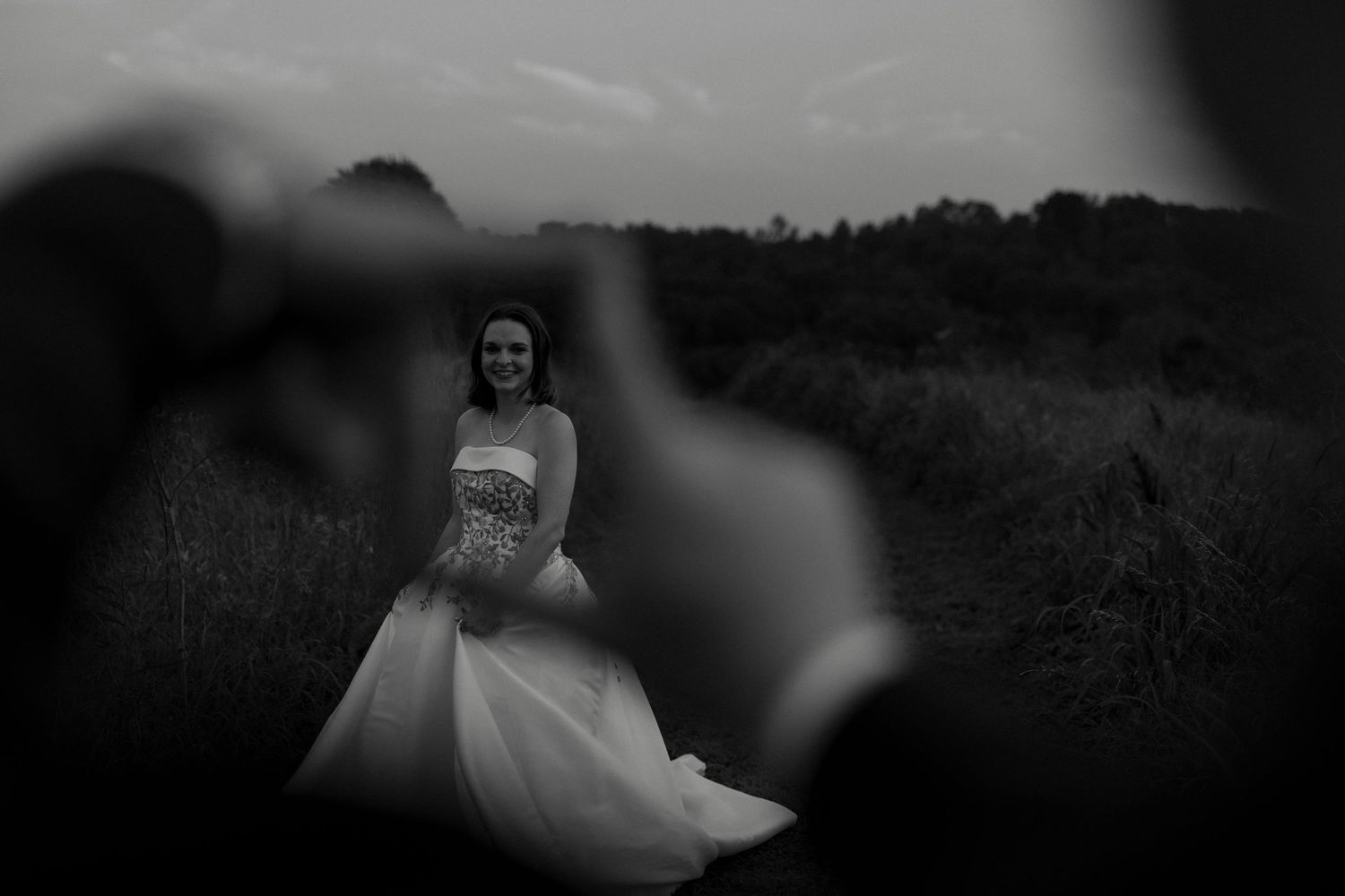 Black and white artistic shot of bride in strapless gown framed by groom's hands outdoors.