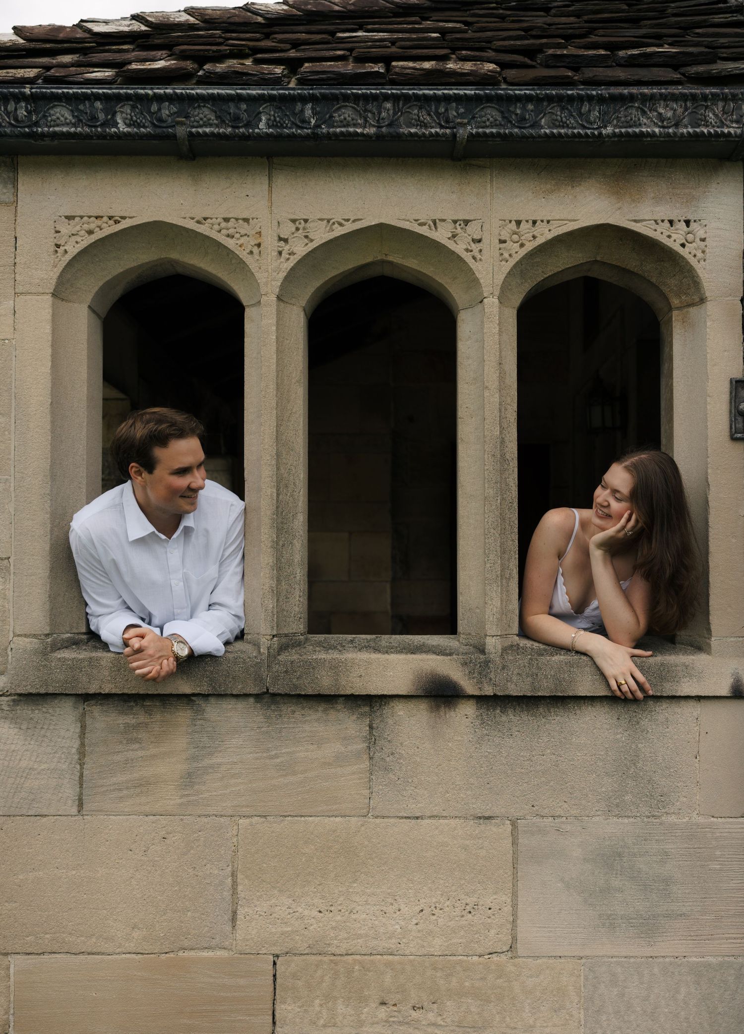 Couple leaning out of ornate gothic stone window arches, sharing a playful romantic moment.
