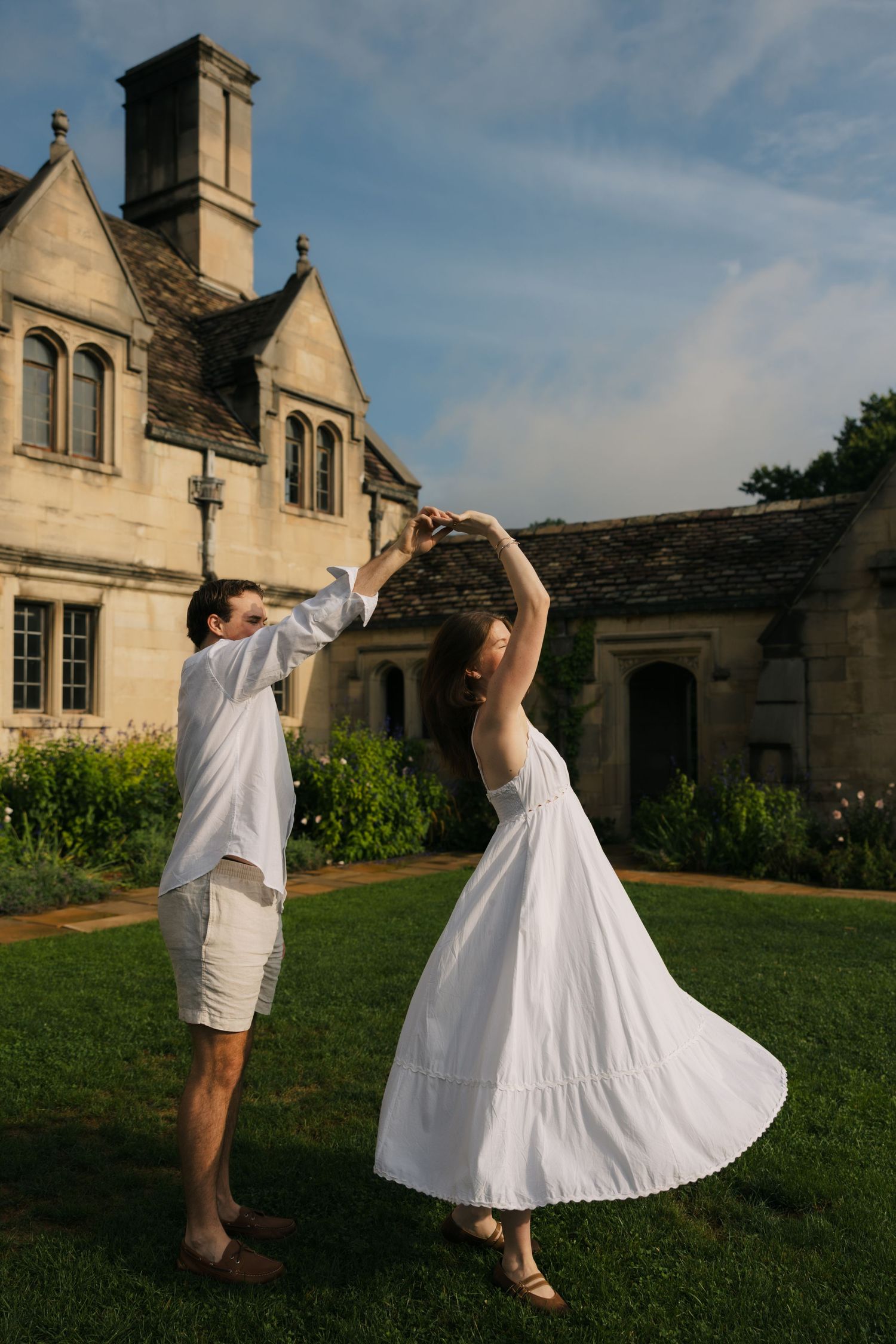 Couple dancing in white outfits on green lawn before historic stone manor house.