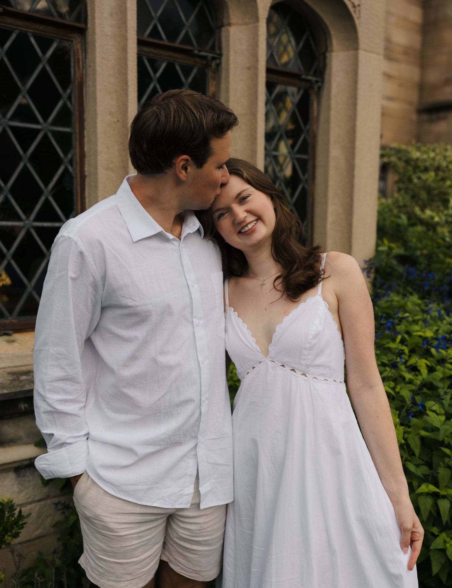 Young couple in white outfits posing before gothic stone building, man kissing womans cheek as she smiles.