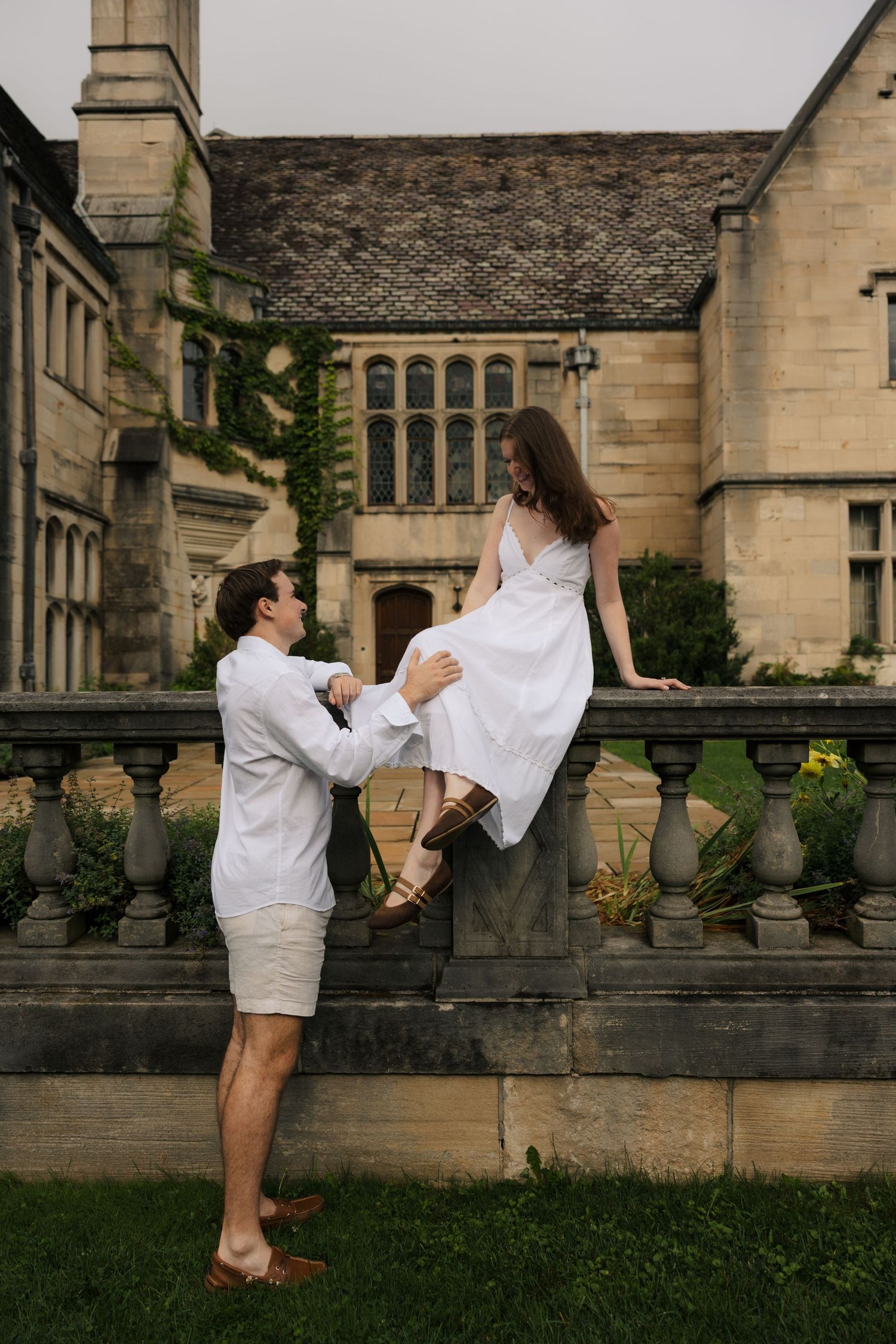 Couple in white outfits posing by stone balustrade of historic manor, man holding woman seated on wall.