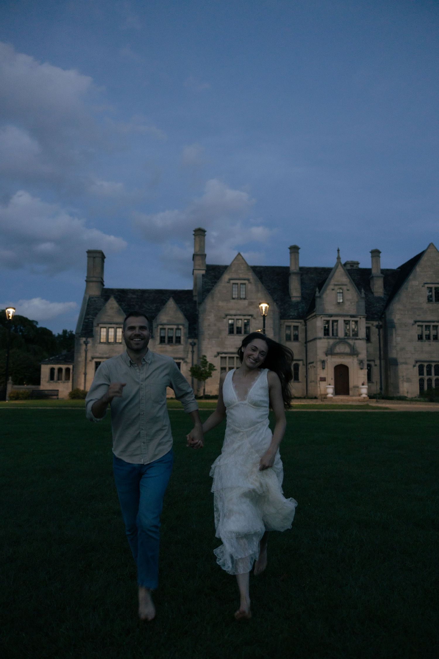 Couple running barefoot hand in hand on lawn before grand stone estate at twilight.
