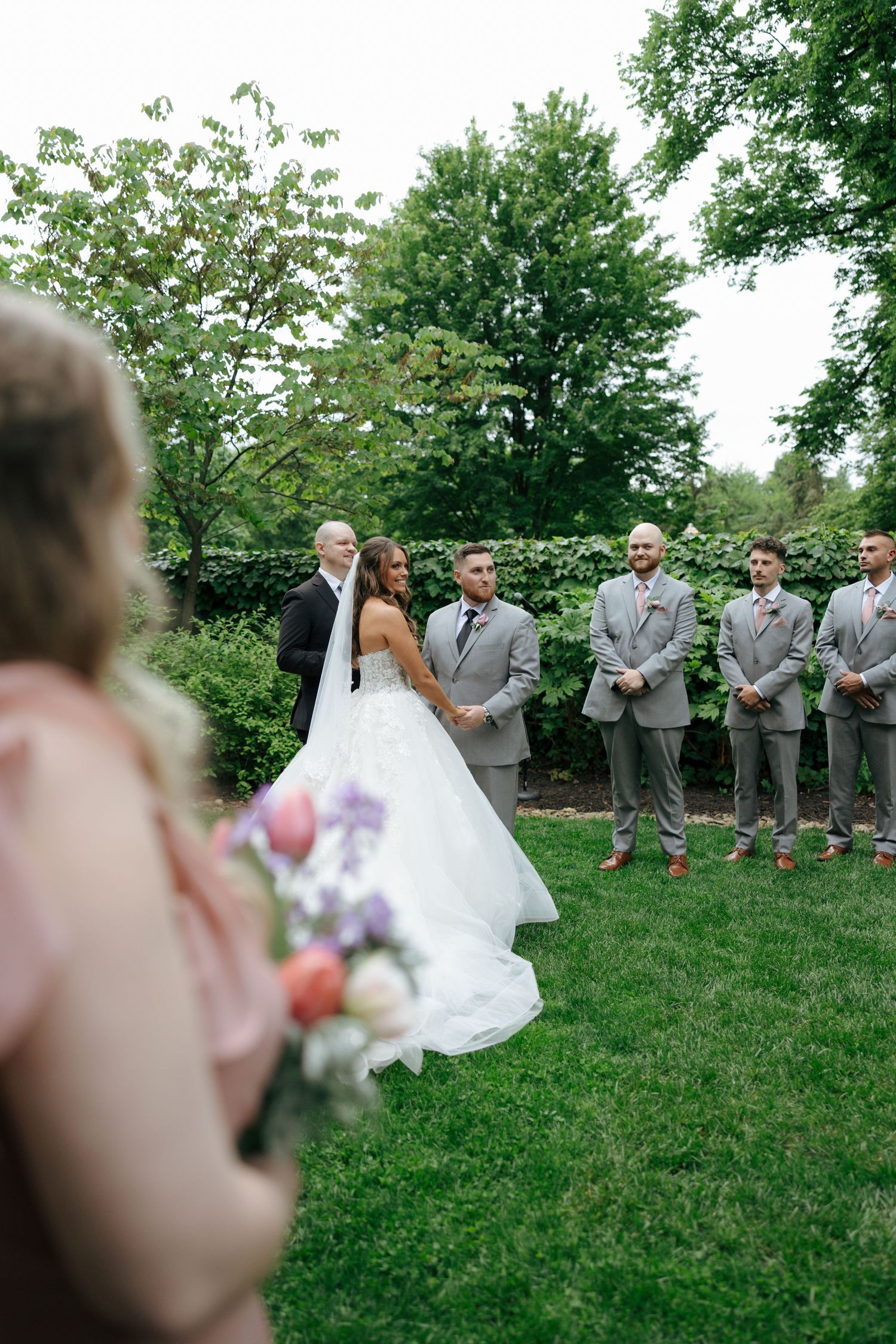 Bride in white ballgown stands at outdoor wedding ceremony with groomsmen in gray suits lined up on green lawn.
