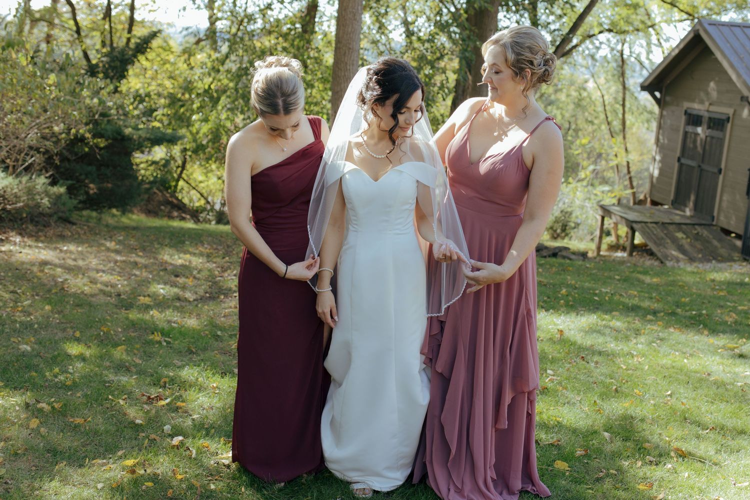 Bride in white gown with veil stands between two bridesmaids in dusty rose and burgundy dresses outdoors.