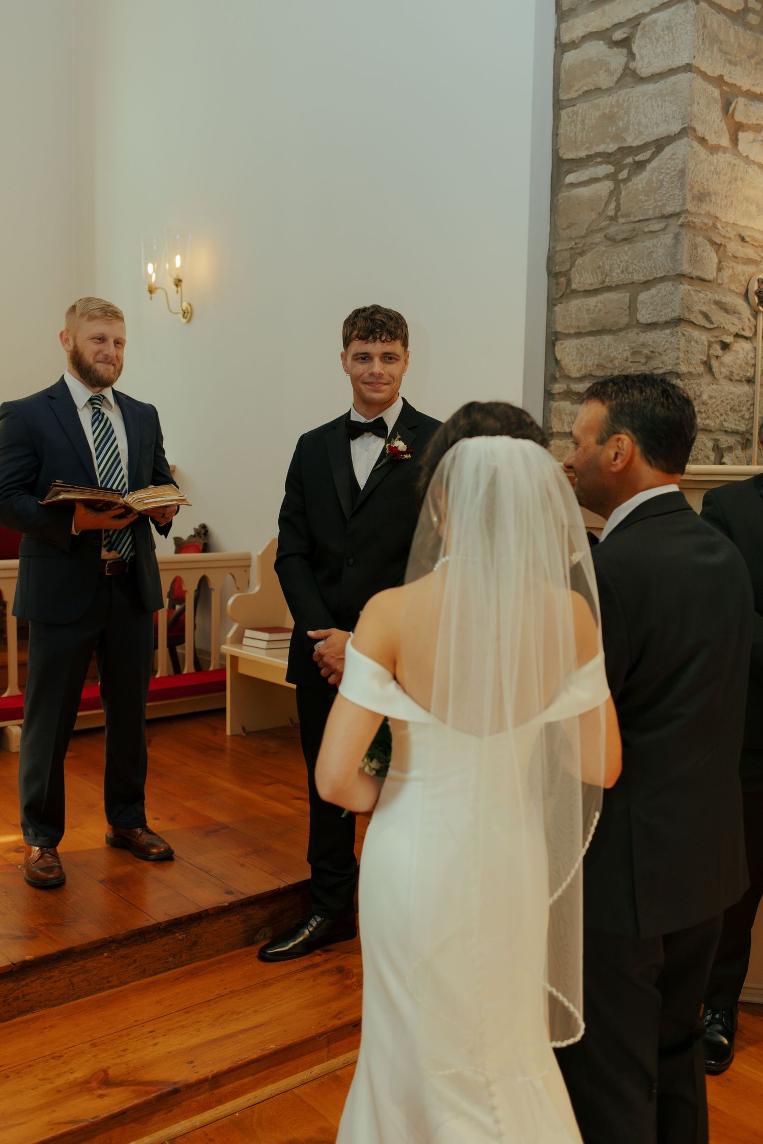 Groom smiling at bride during wedding ceremony in a stone church with officiant holding a book.