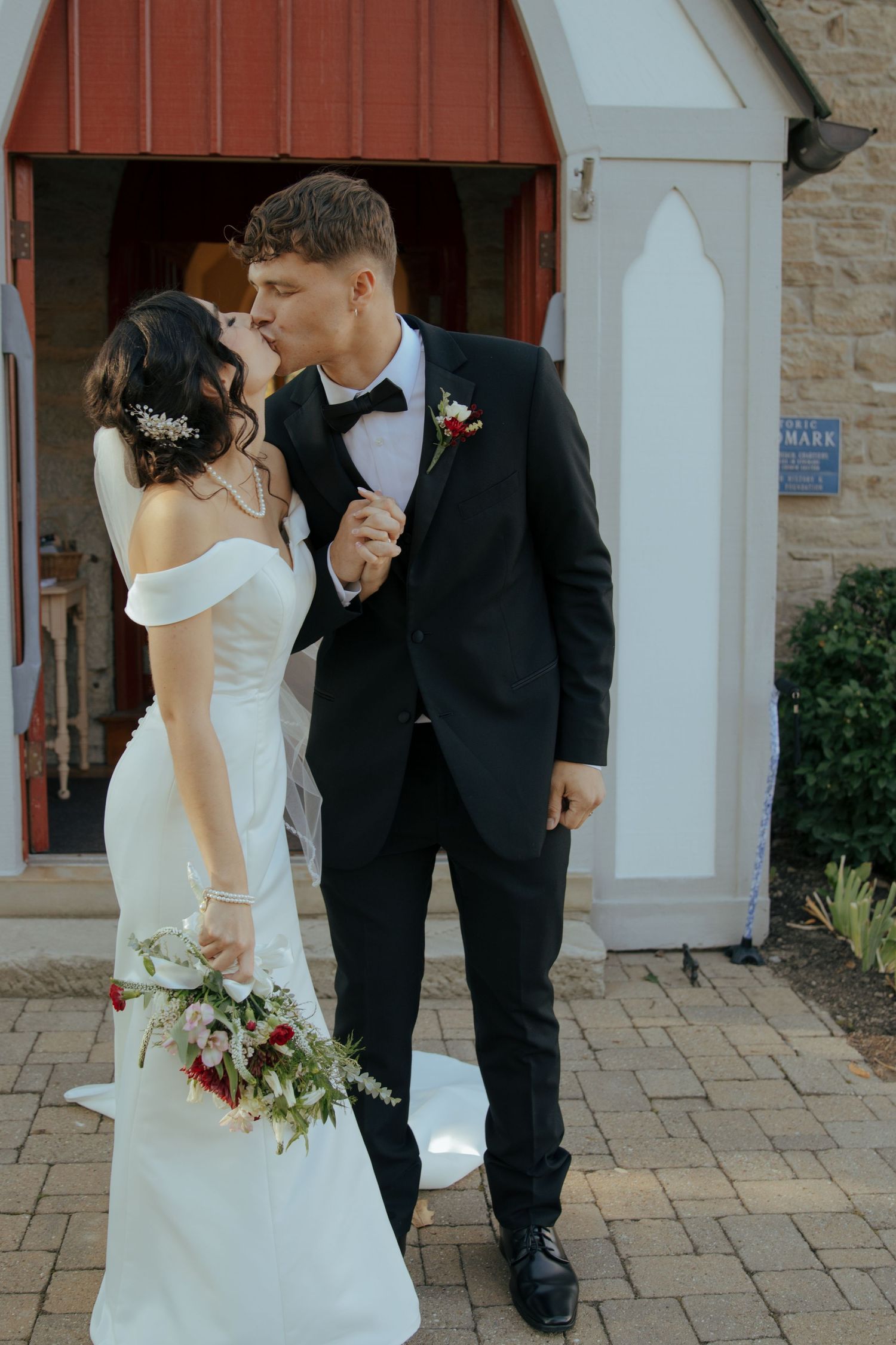 Bride and groom sharing a kiss outside a historic stone chapel, bride holding a red and white bouquet.