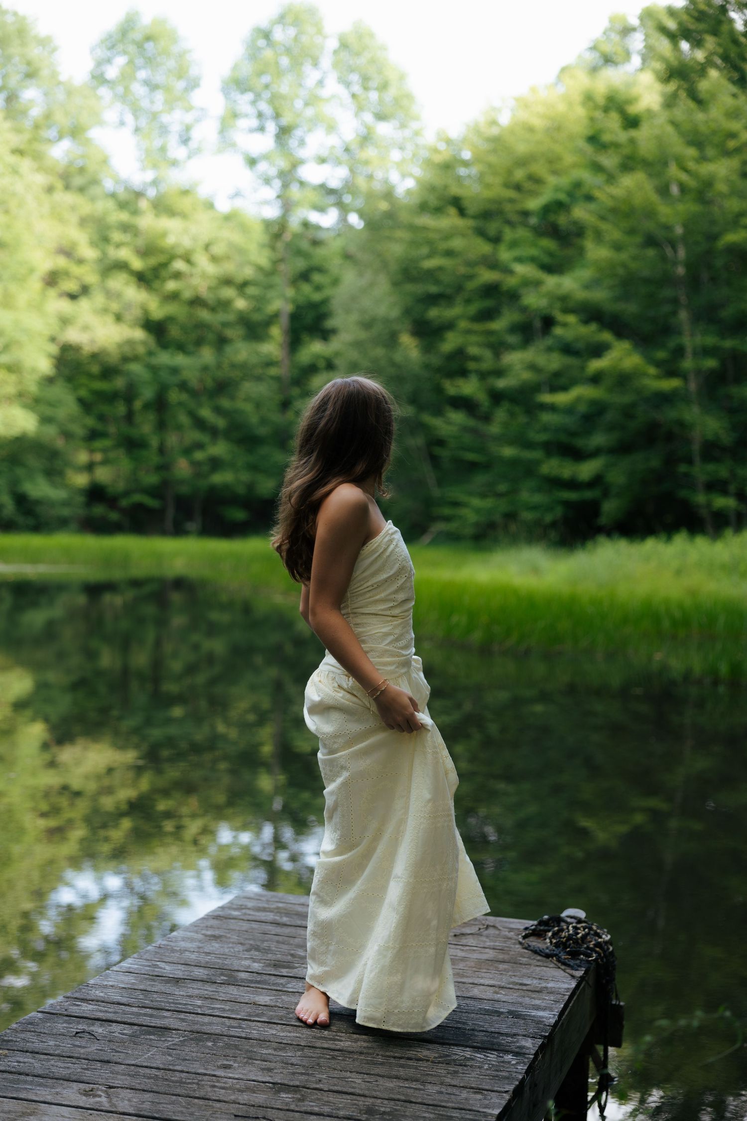 Young woman in white strapless dress standing barefoot on rustic wooden dock by serene forest lake.