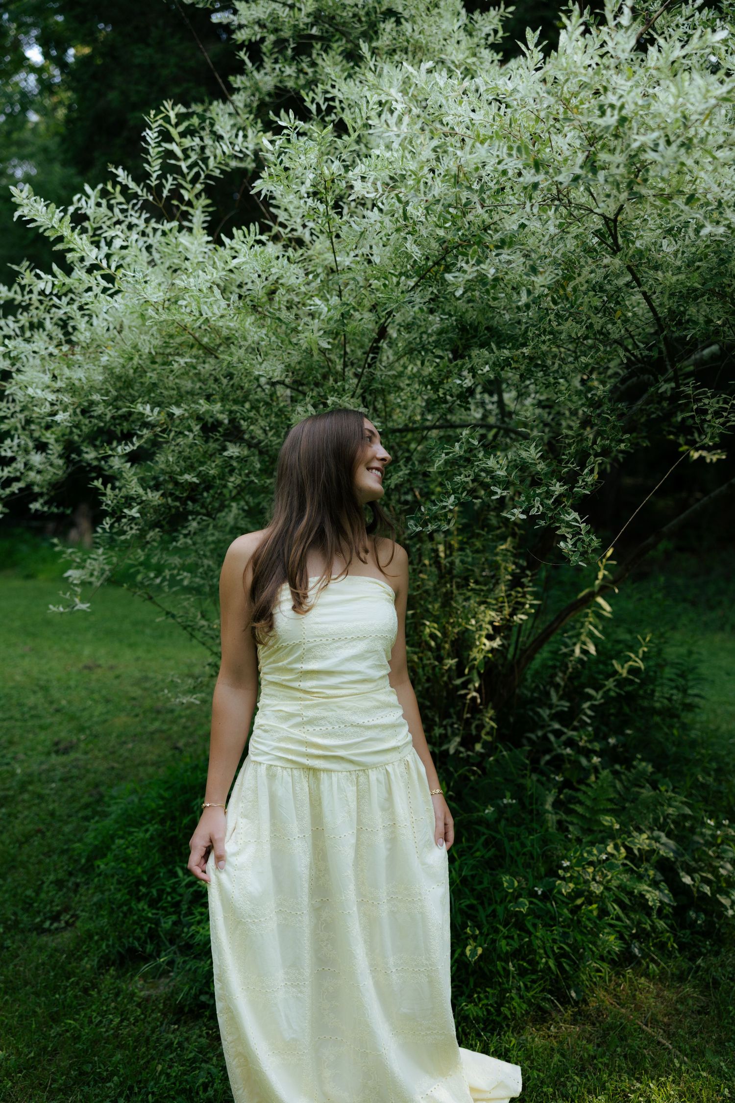 Young woman in a pale yellow strapless maxi dress standing in front of white blooming bushes outdoors.