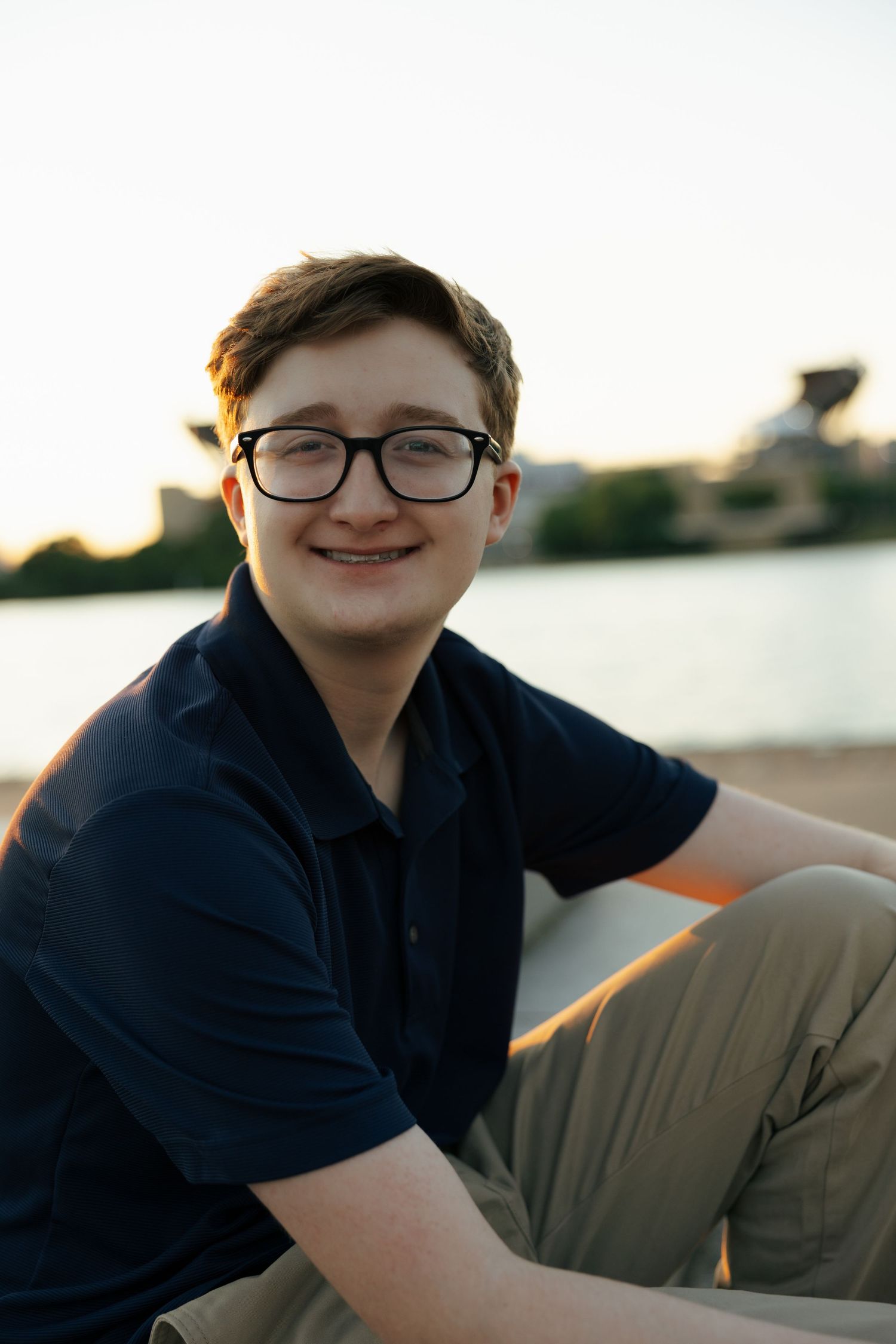 Smiling teen in glasses and navy polo sitting outdoors near a lake at dusk.