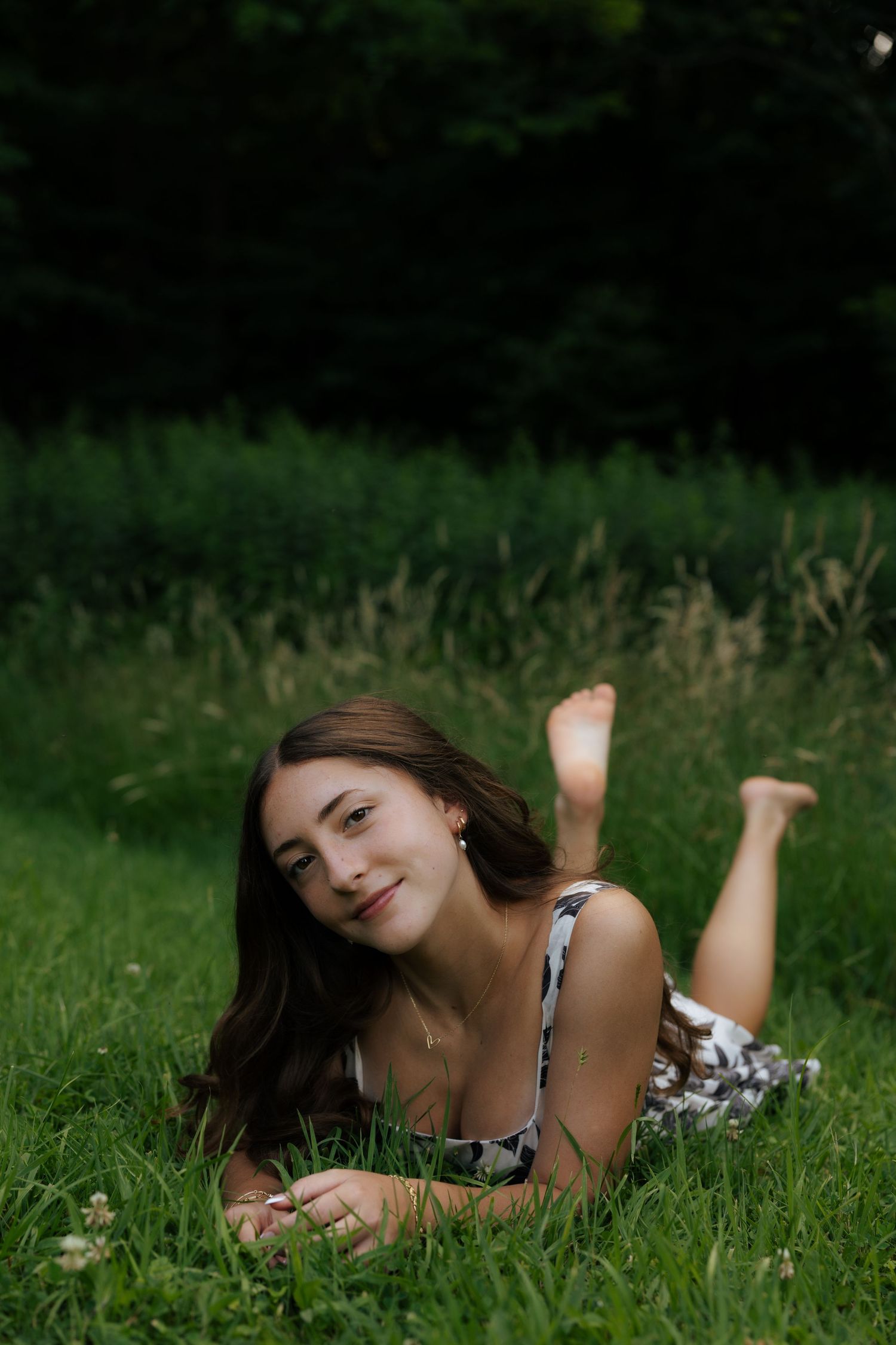 Young woman in floral dress lying on green grass smiling, barefoot with trees in background.
