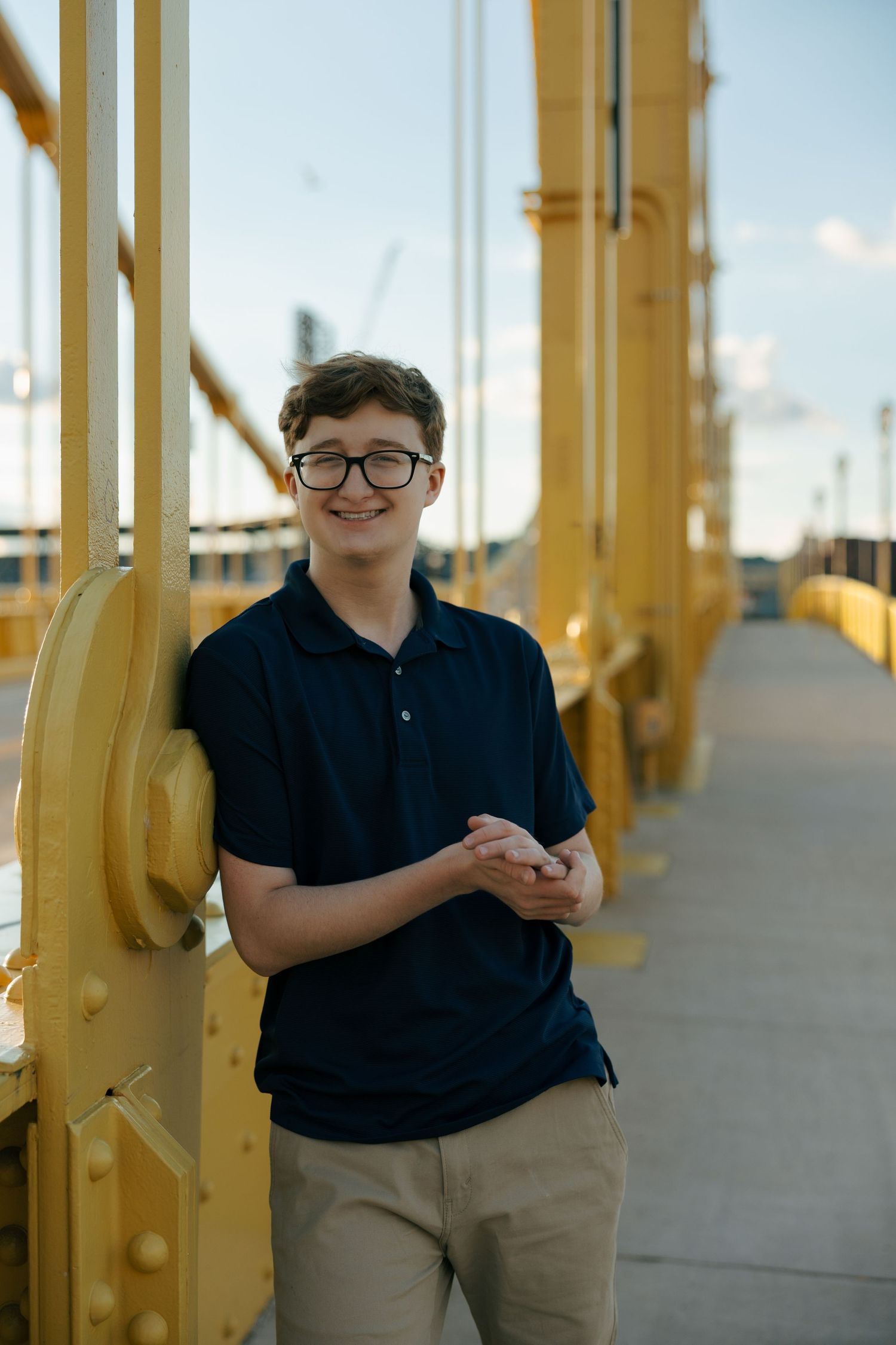 Young teen boy with braces and glasses smiles while leaning against yellow bridge structure outdoors.