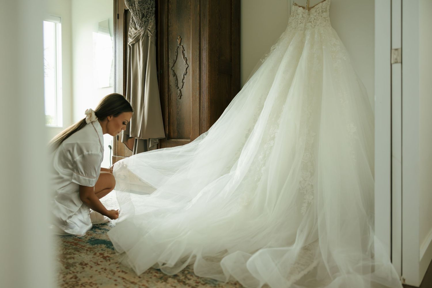 A person carefully arranges a flowing white wedding gown in an elegant room with soft natural light.