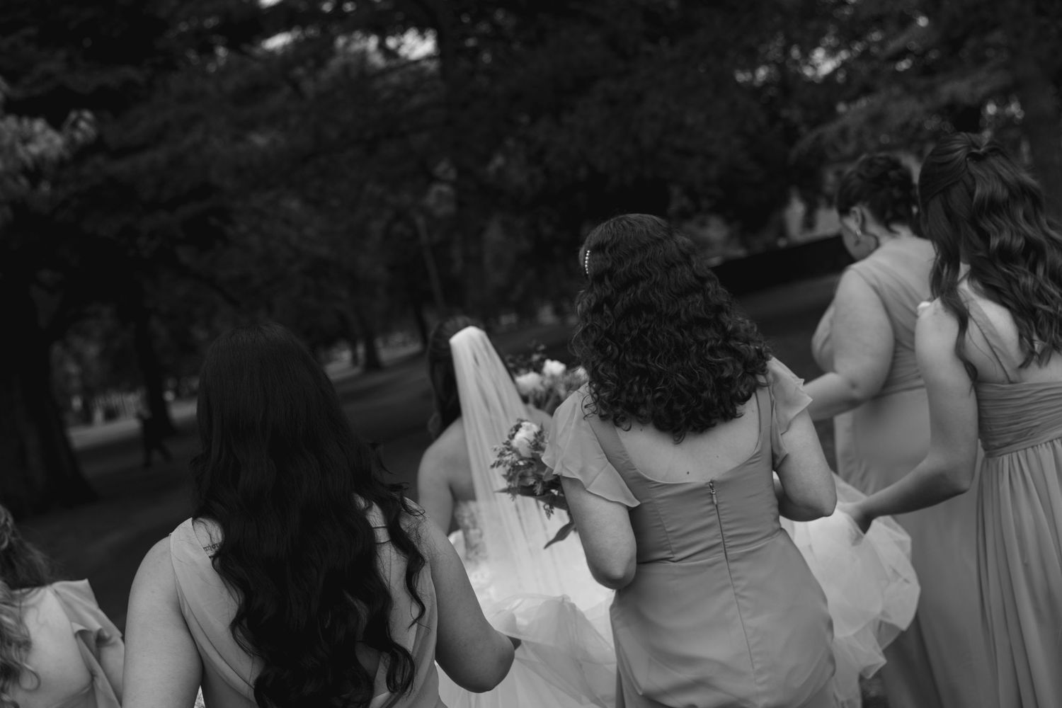 Black and white photo of bride and bridesmaids walking together outdoors at wedding celebration.