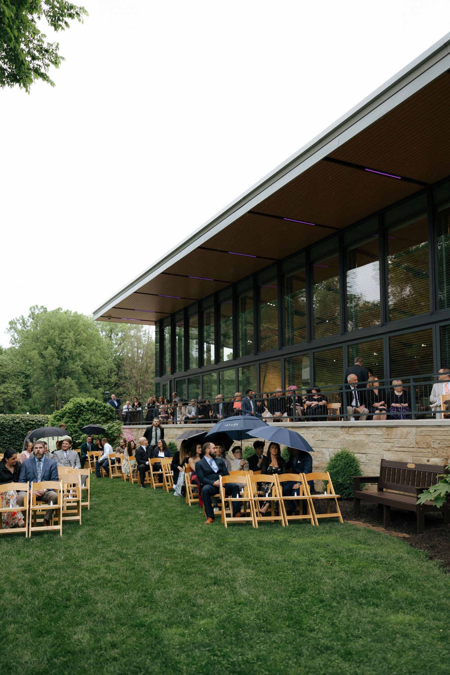 Outdoor wedding ceremony on green lawn beside modern glass building with guests seated in wooden chairs.