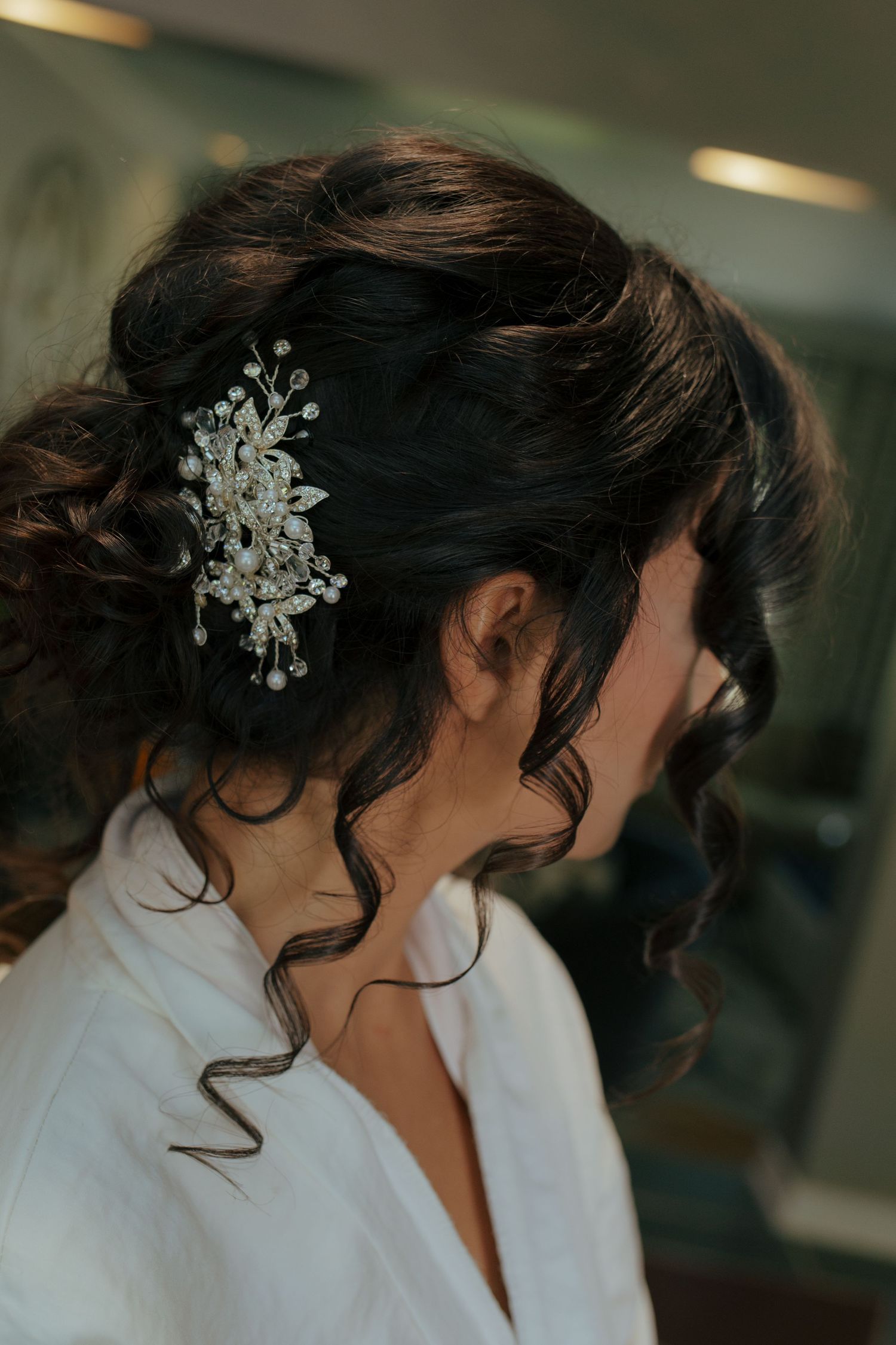 Bride with dark curly updo adorned with a delicate pearl and crystal hair comb, wearing a white robe.