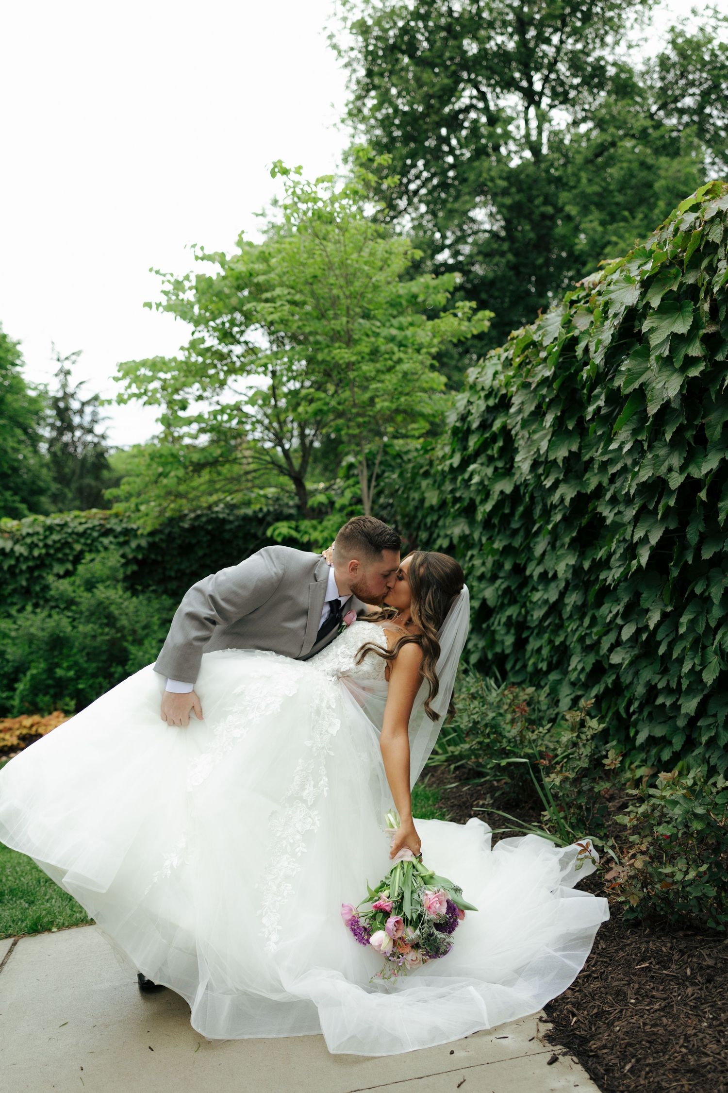 Bride and groom share a romantic dip kiss in a lush garden, bride holding a pink bouquet in a flowing white gown.
