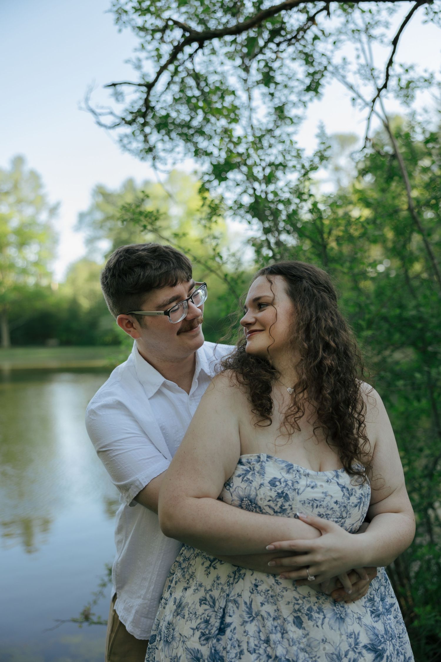 Couple embracing by a pond, man in white shirt hugging woman in blue floral dress, surrounded by lush green trees.