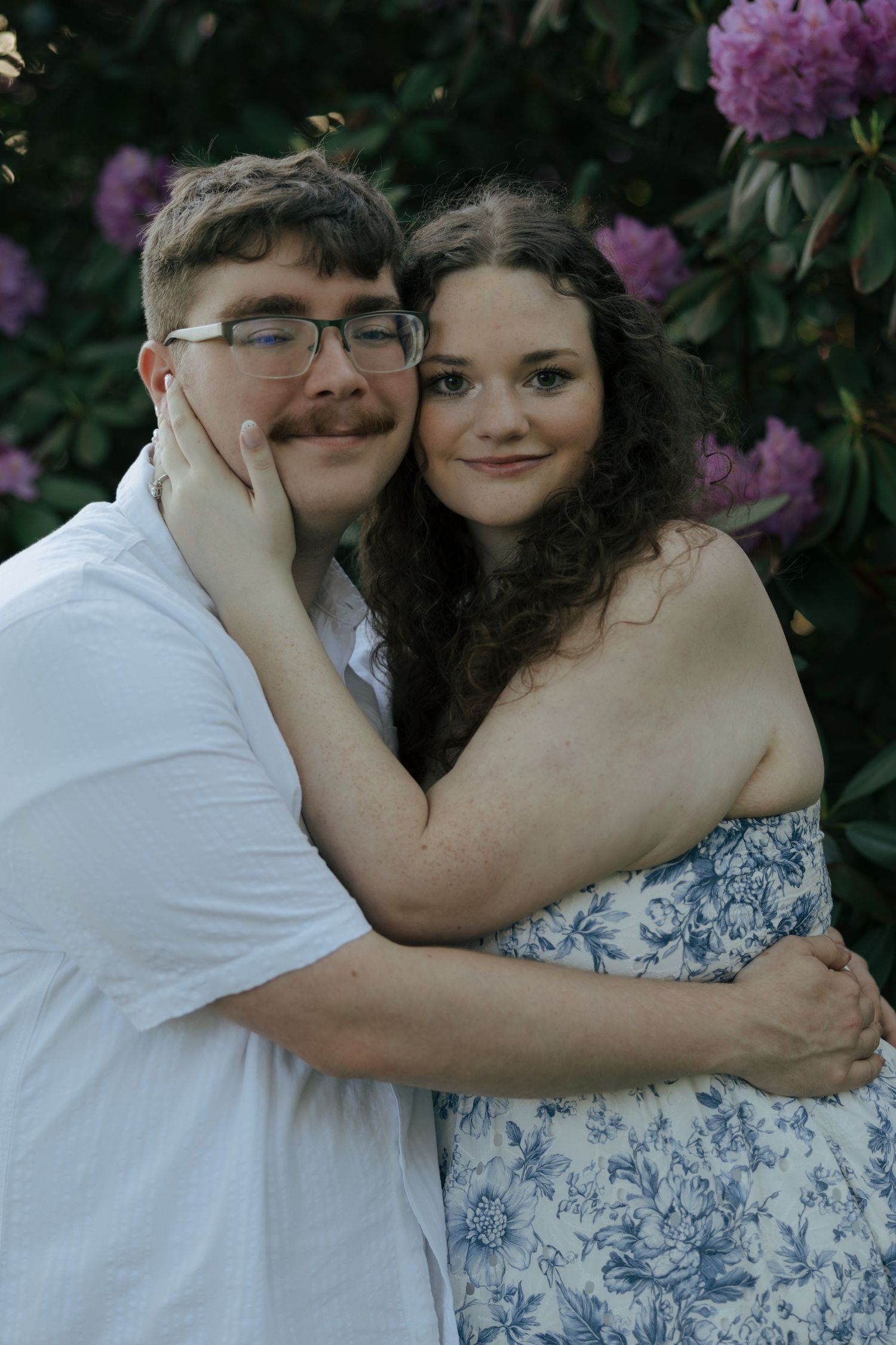 Happy couple hugging closely in front of vibrant purple rhododendron flowers, woman cradling man's face.