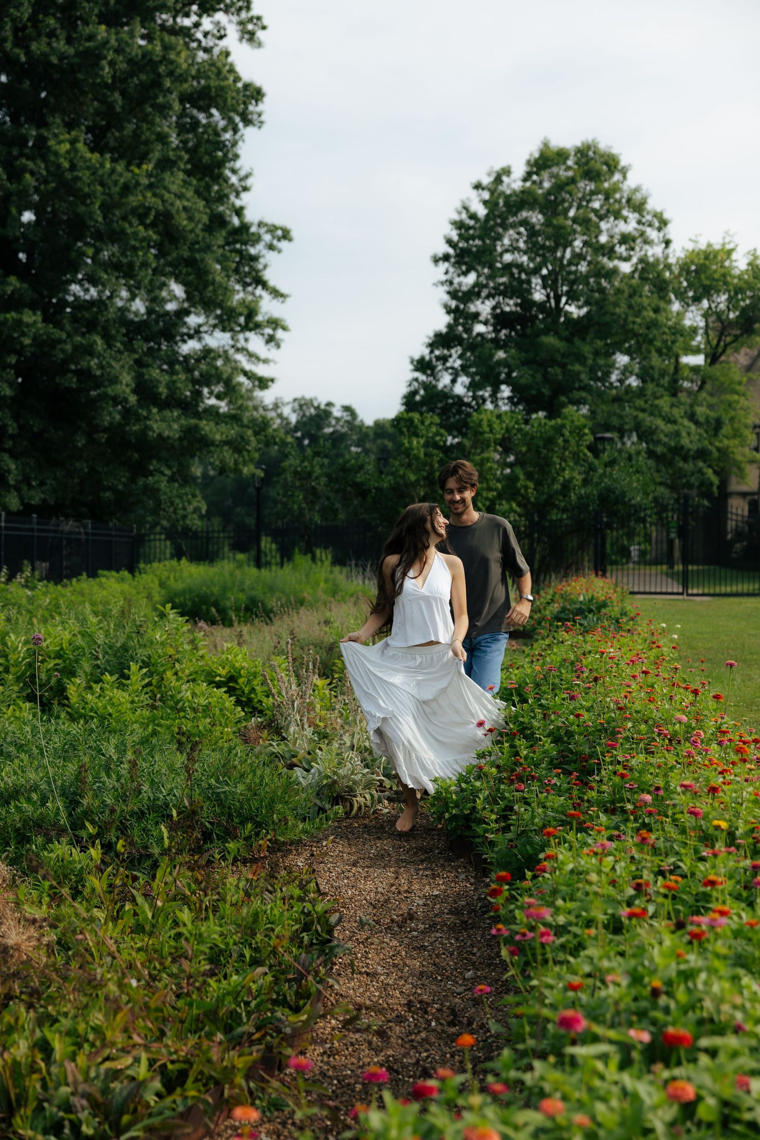A couple walks hand in hand along a flower-lined garden path surrounded by trees.