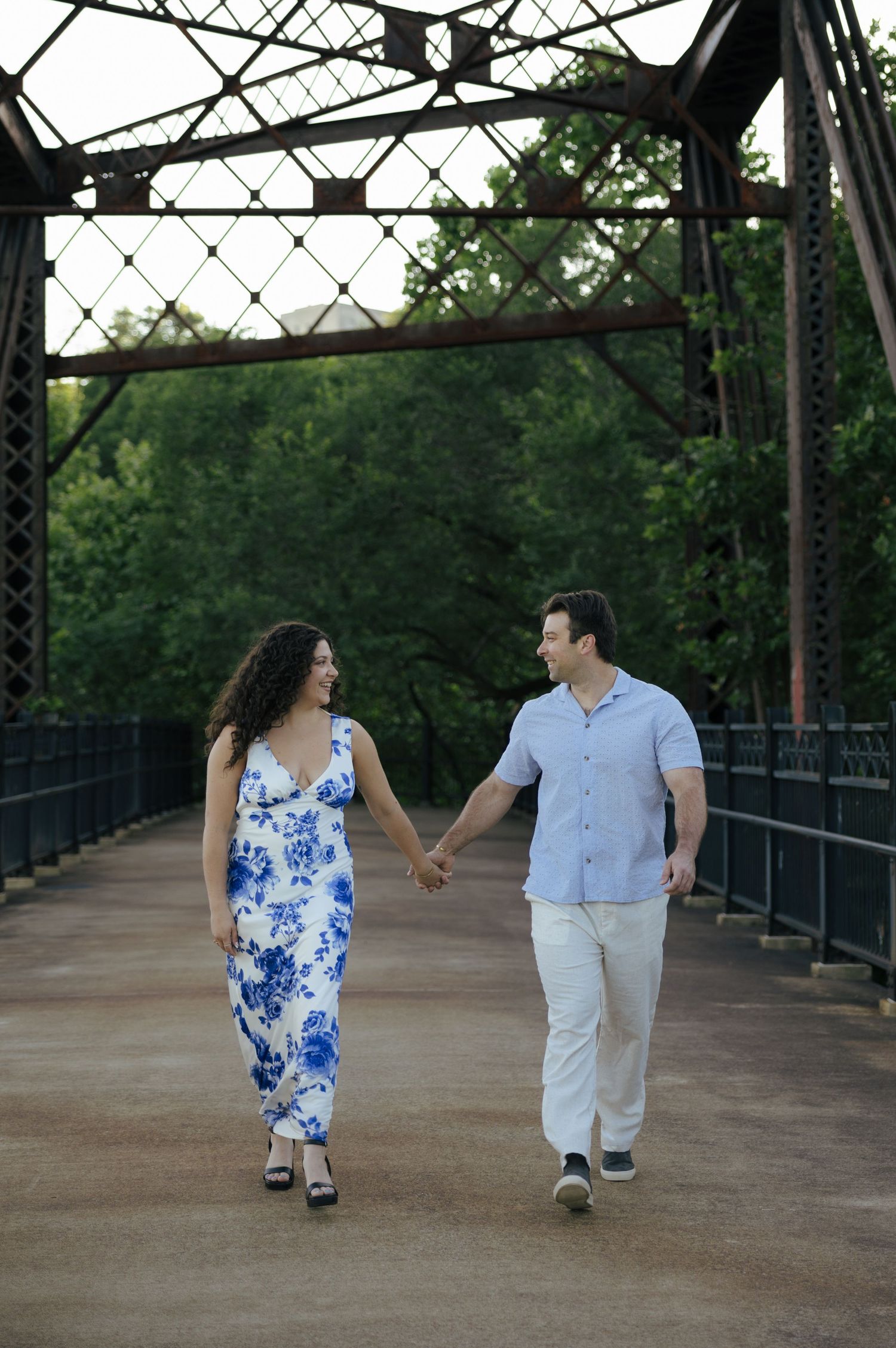 Engaged couple walking hand-in-hand across historic iron bridge, woman in blue floral dress.