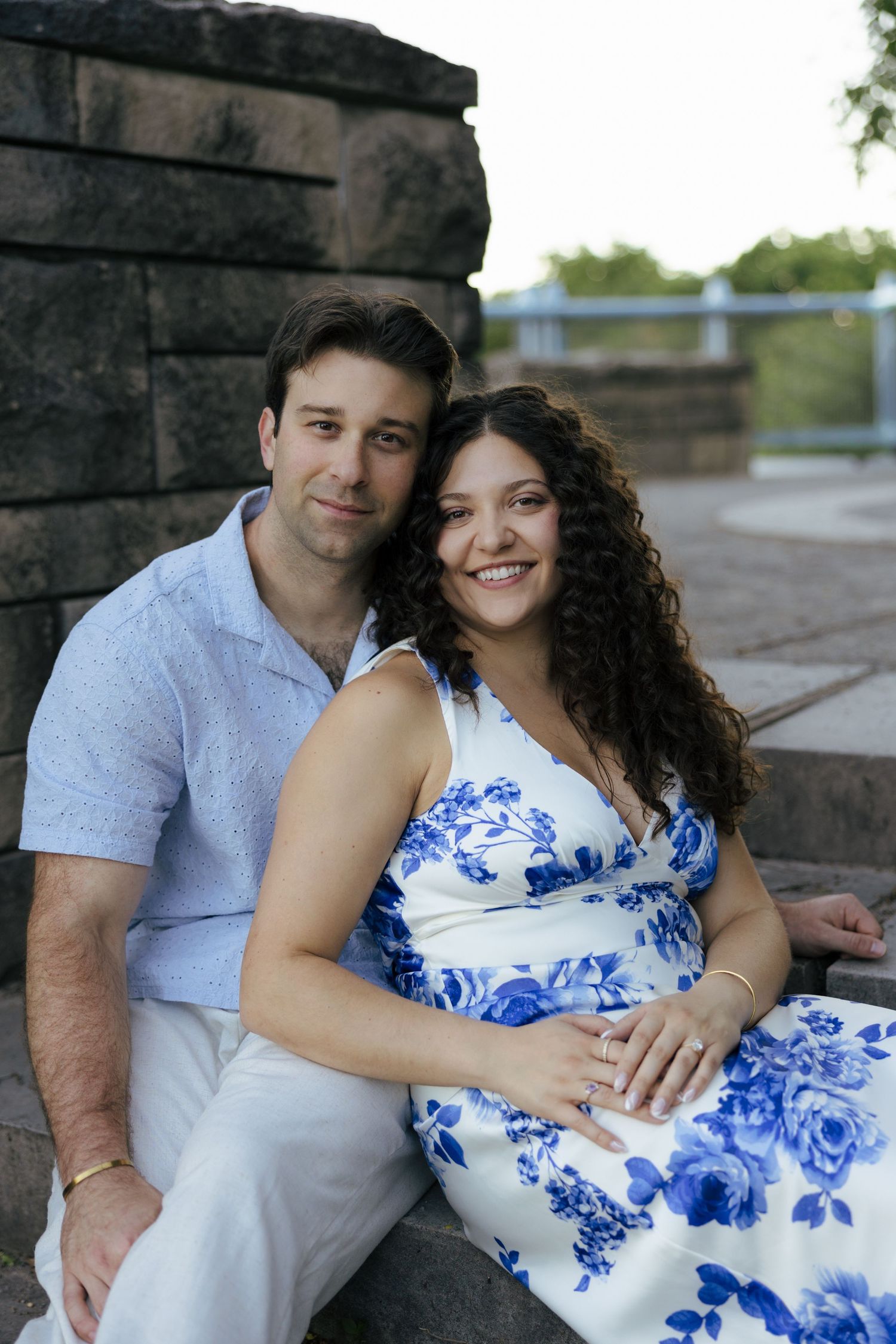 A happy engaged couple sits together on stone steps, woman in a blue floral dress showing her ring.