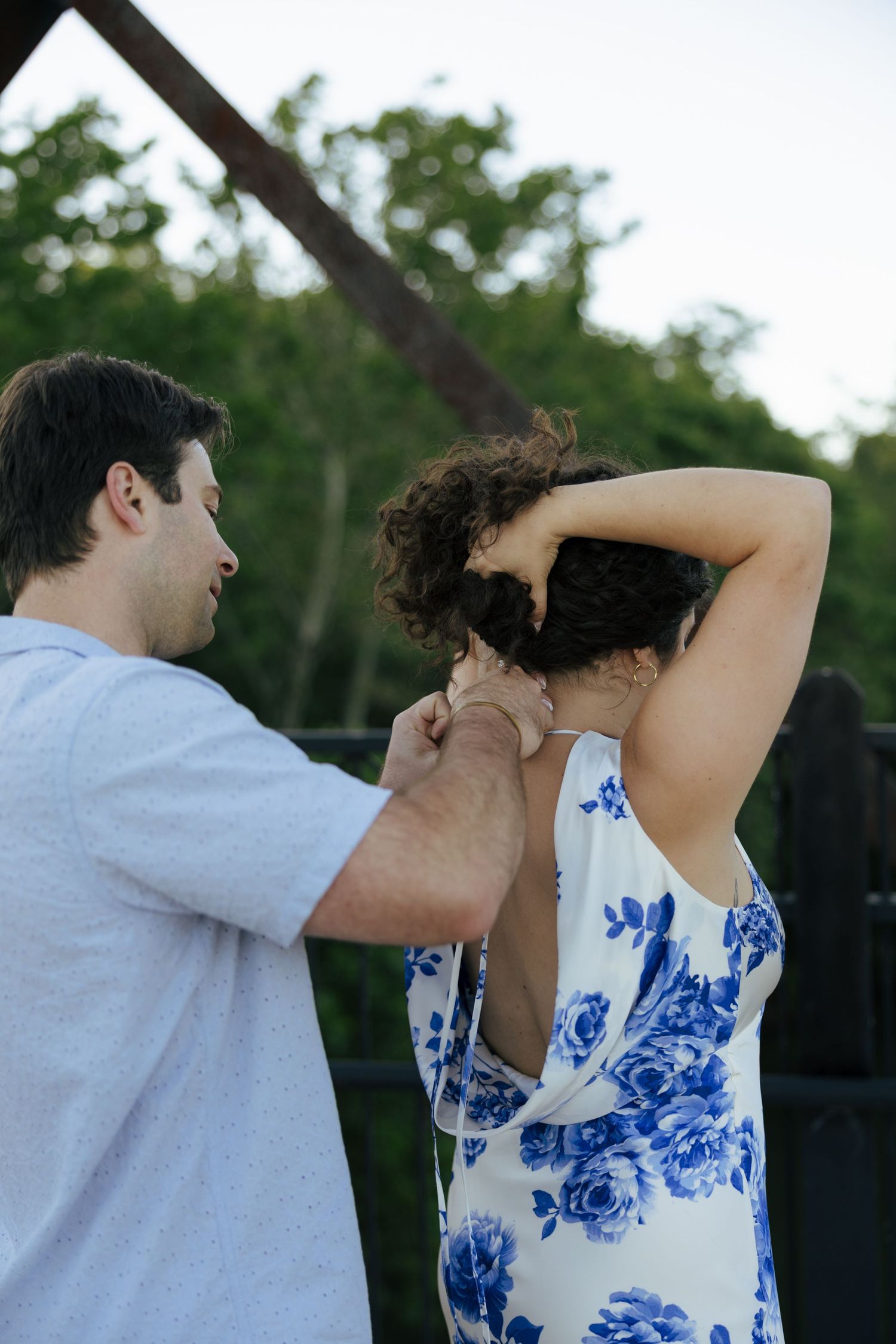 A man carefully fastens a necklace around a woman's neck outdoors near an iron bridge structure.