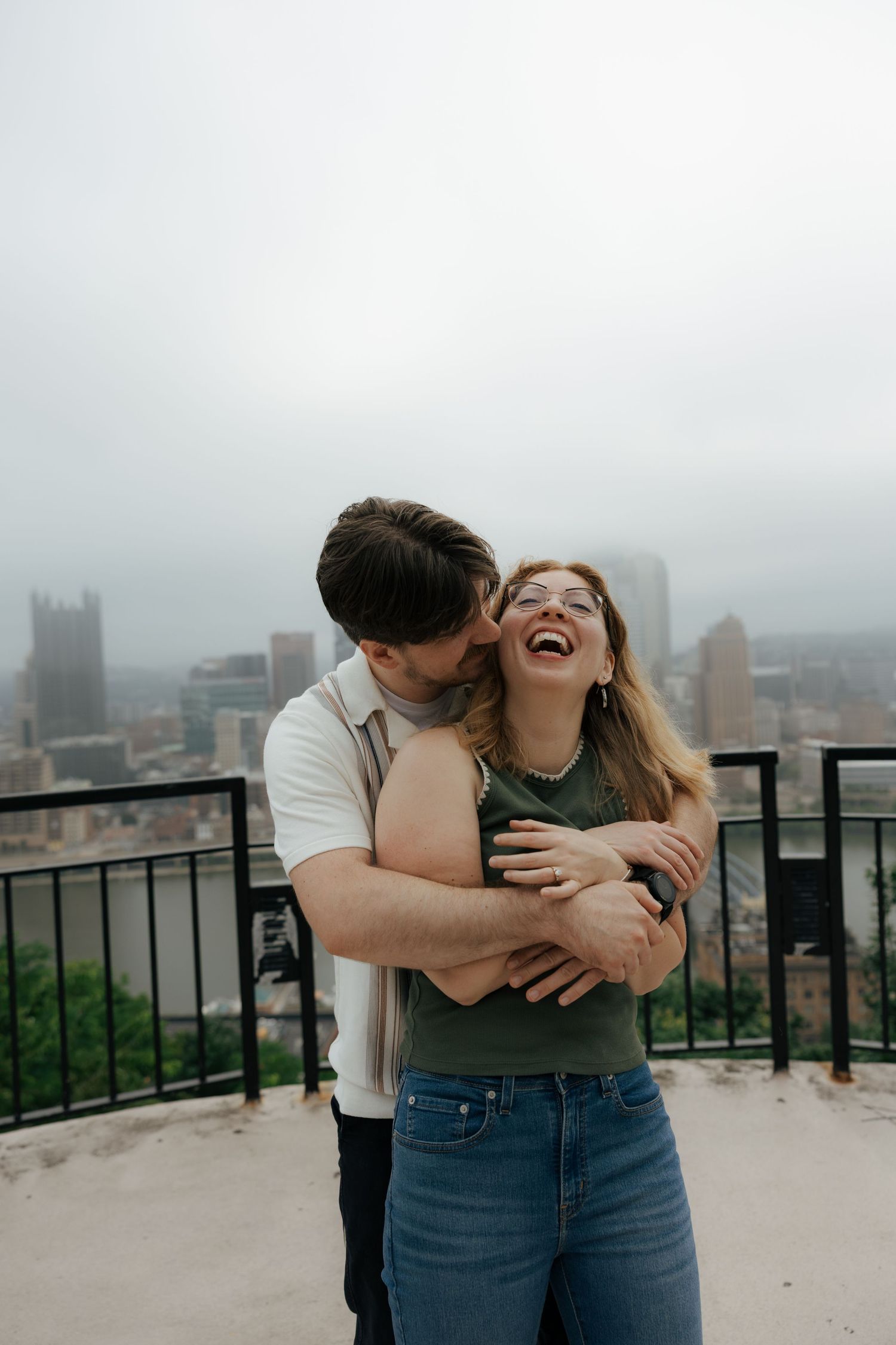 A joyful woman laughs as her partner embraces and kisses her cheek with a foggy city skyline behind them.
