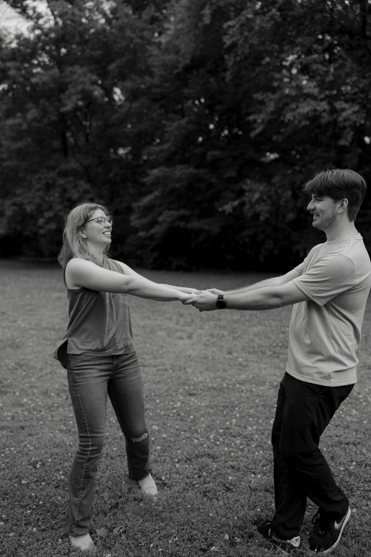 Black and white photo of a laughing couple holding hands and spinning in a grassy park.