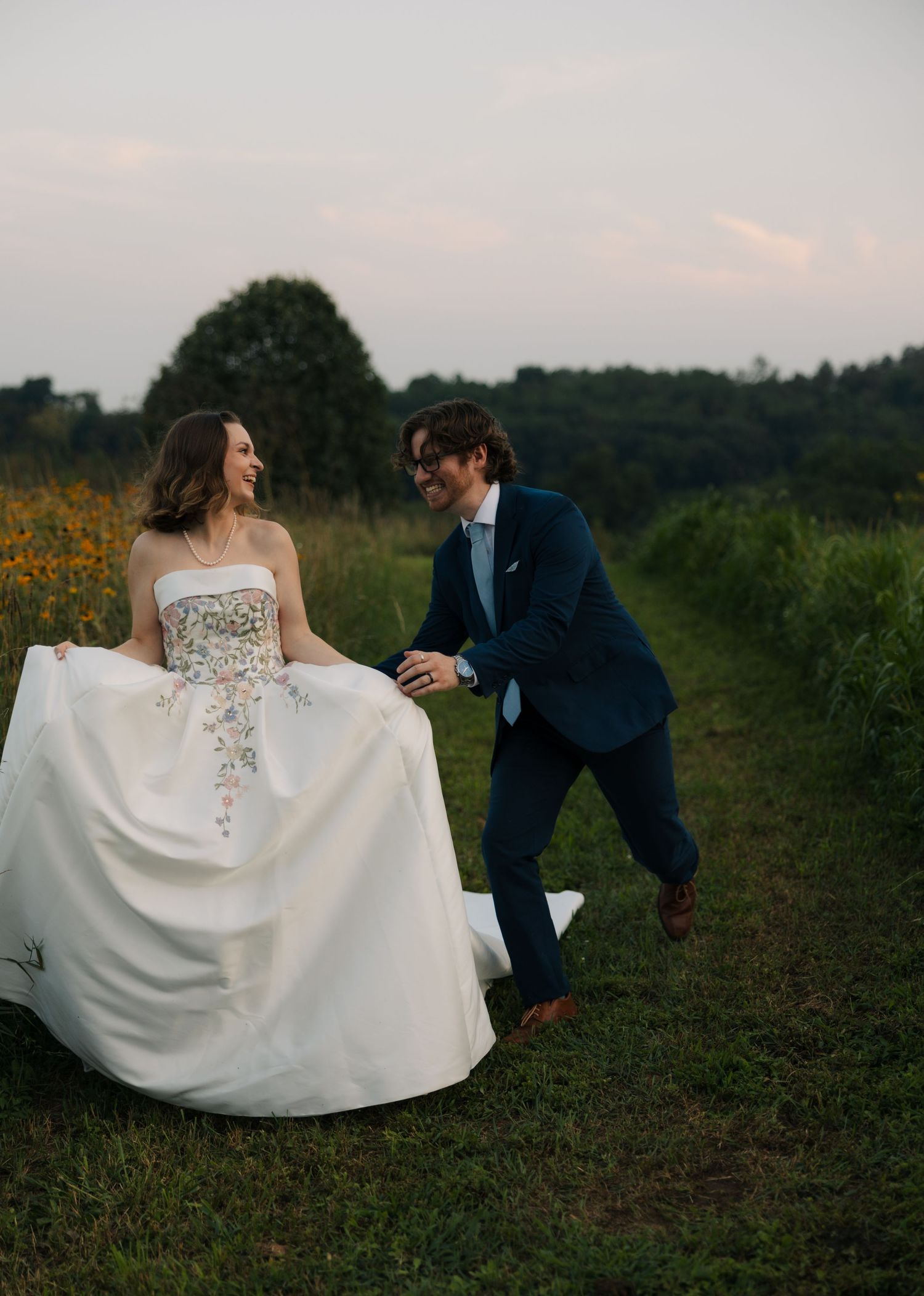Laughing bride and groom hold hands while walking playfully along a wildflower meadow path at sunset.