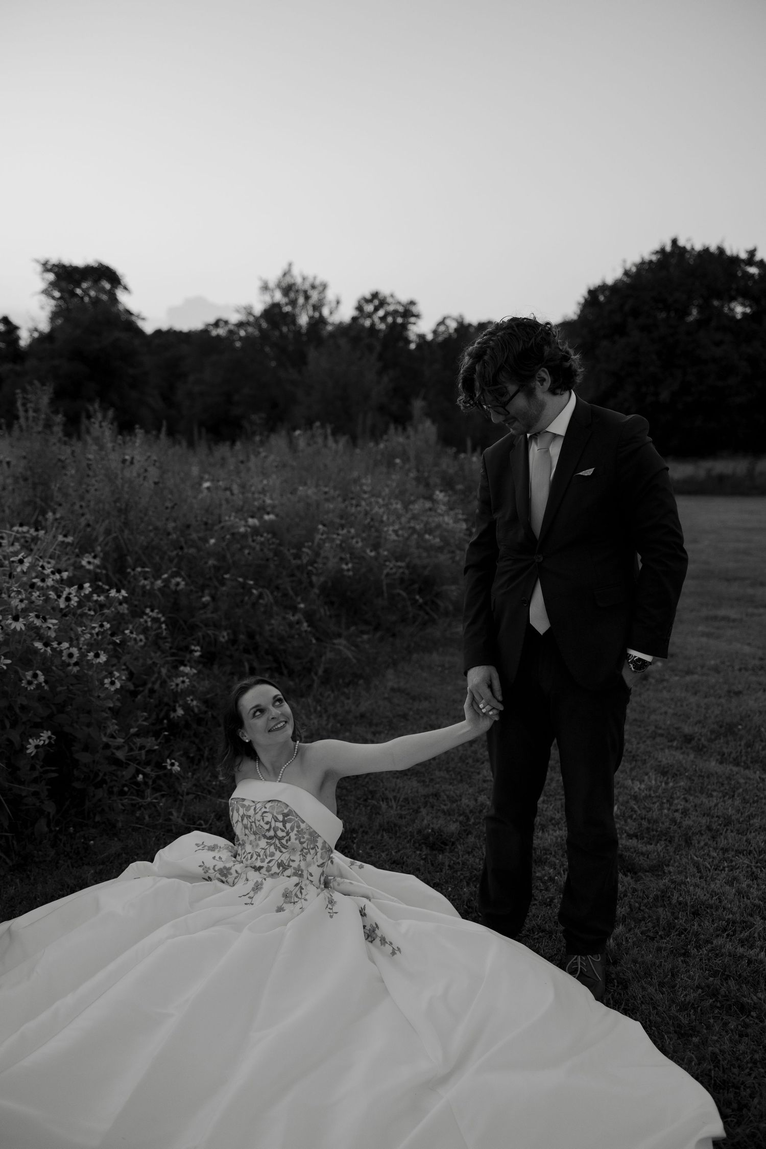 Black and white photo of bride sitting on grass reaching up to hold groom's hand at twilight.