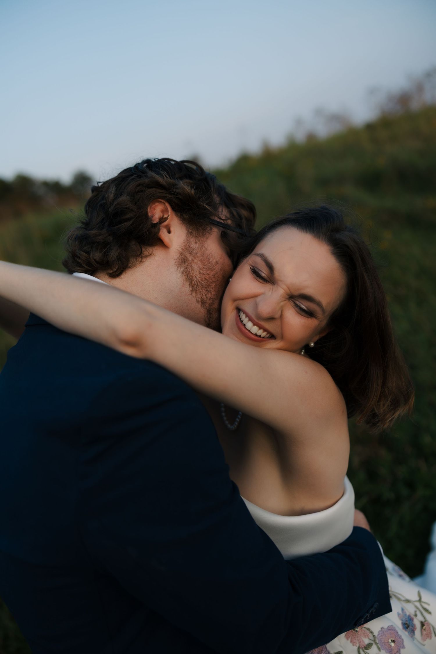 Joyful bride laughs while embracing groom in navy suit during outdoor golden hour shoot.