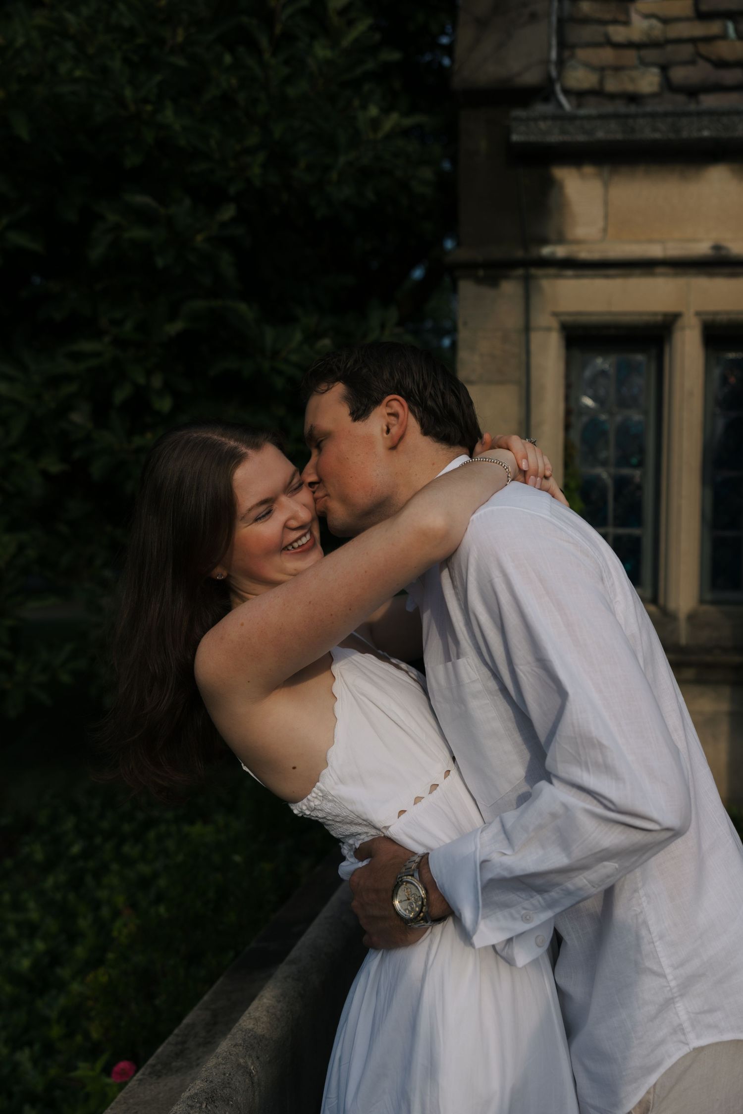 Romantic couple in white outfits sharing a tender kiss outdoors beside a stone manor building.