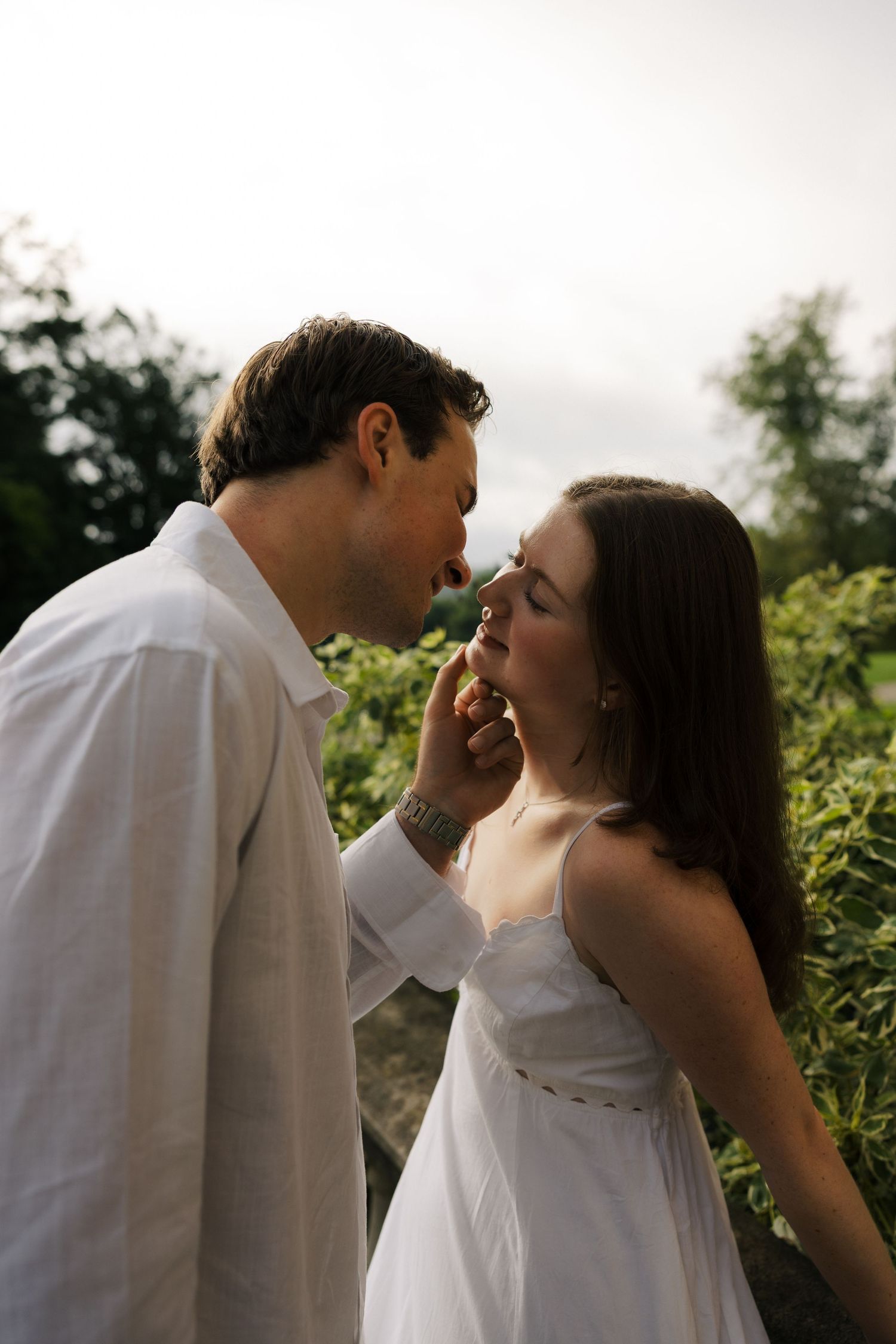 A couple in white outfits share a tender moment outdoors, the man gently touching the woman's face.