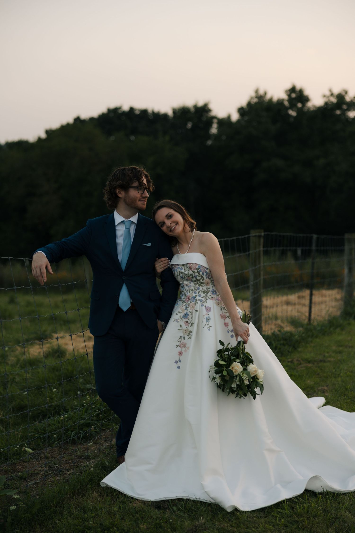 Bride in strapless floral wedding gown holds bouquet beside groom in navy suit at dusk by fence.