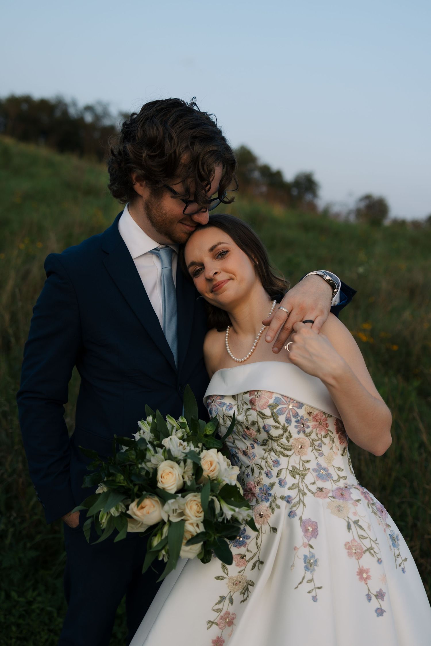 Groom in navy suit kisses brides head as she leans back holding white floral bouquet in green meadow.