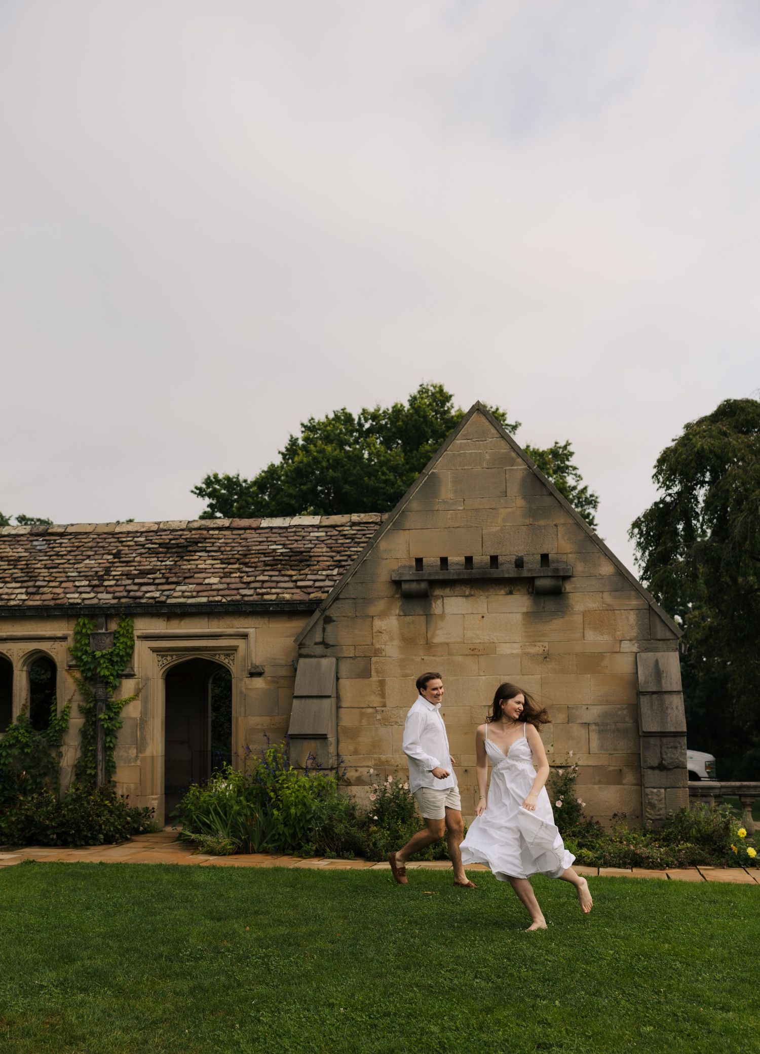 Couple in white outfits running and laughing across green lawn beside a historic stone cottage.