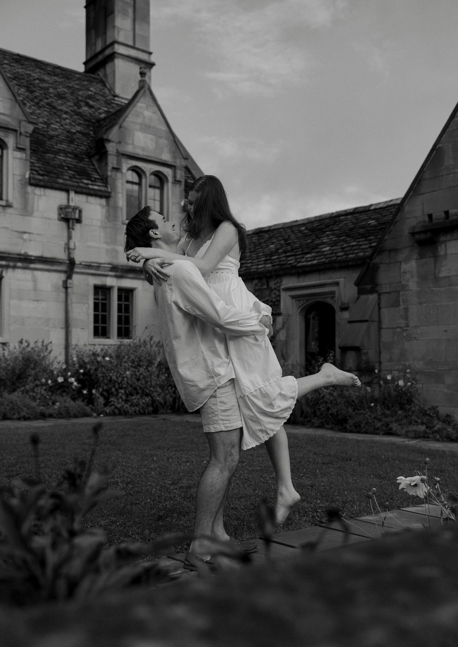 Couple embraces joyfully outside historic stone estate in black and white engagement photo.