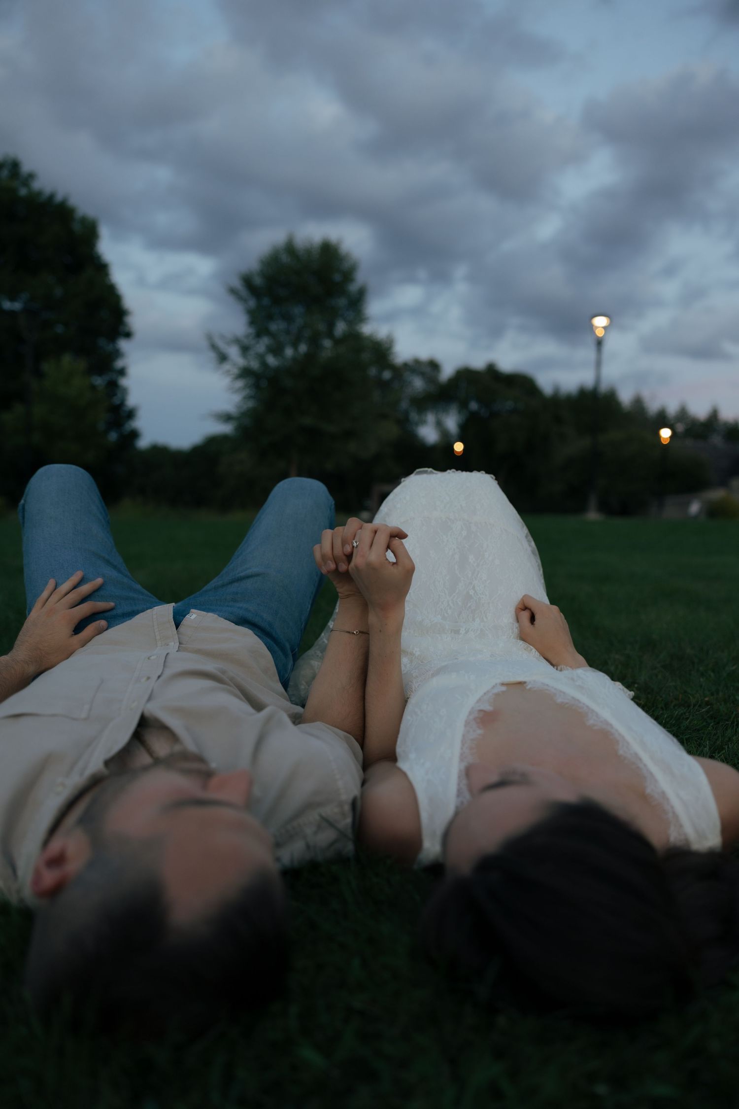 Couple holding hands while lying on green grass at dusk under a cloudy sky.