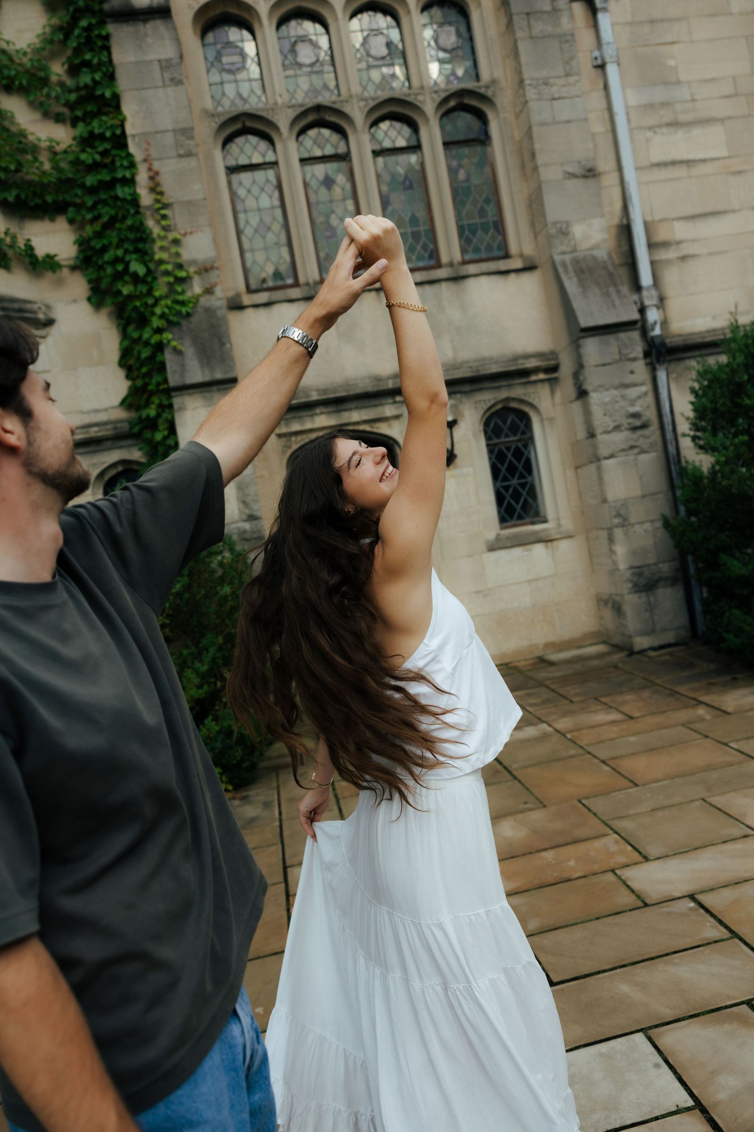 Man spinning woman in white dress outdoors near a stone building covered in ivy.