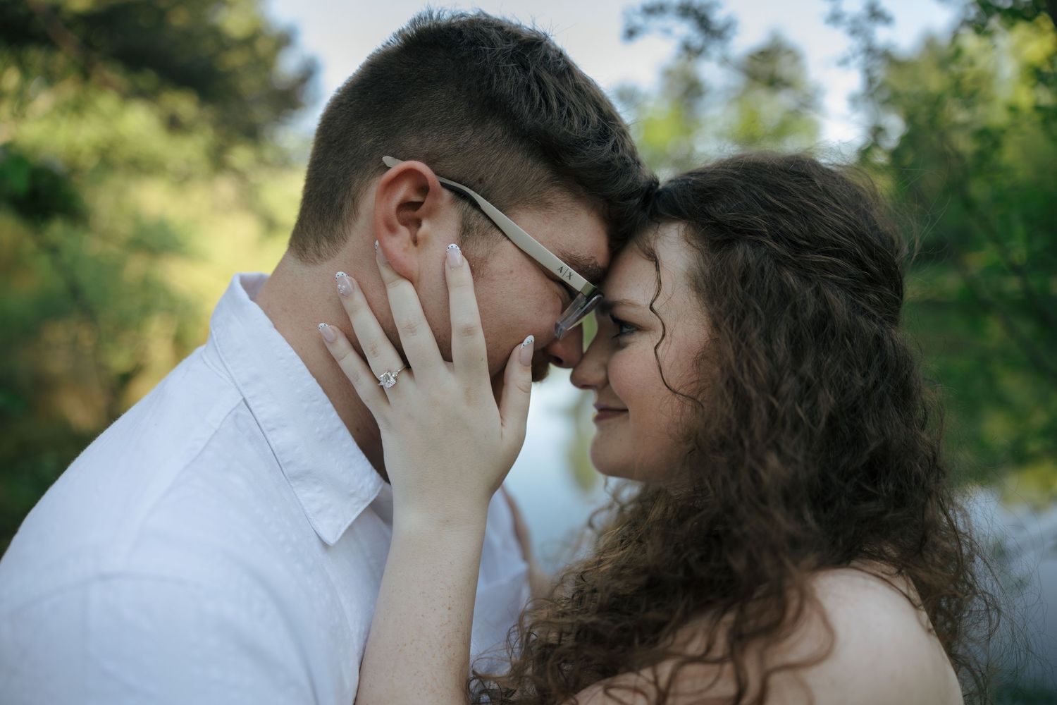 Romantic couple touching foreheads tenderly outdoors, woman wearing engagement ring, sharing intimate moment in nature.