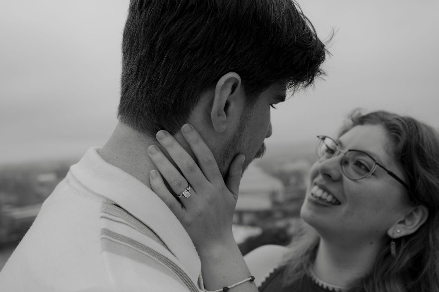 Black and white photo of a smiling couple sharing a romantic moment, woman wearing glasses and engagement ring outdoors.