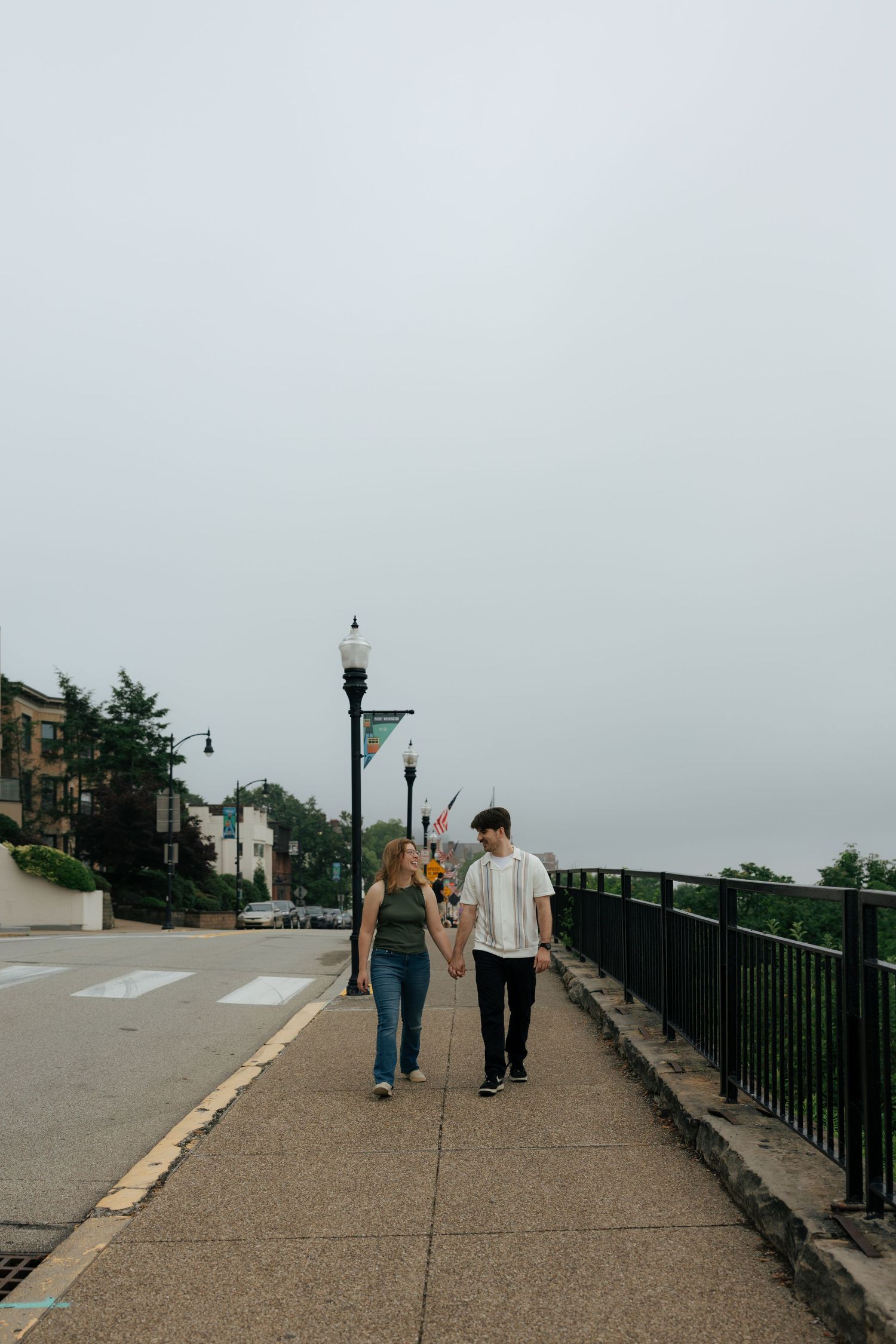 Young couple holding hands while strolling along a scenic urban walkway with black iron railings on an overcast day.