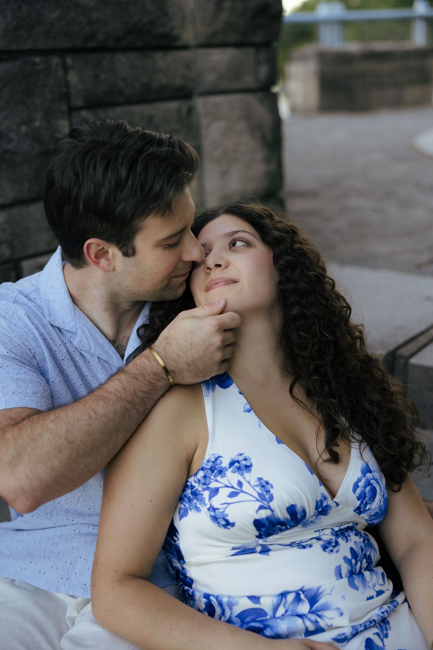 Man gently tilting woman's chin as she gazes up at him while seated outdoors near stone wall.