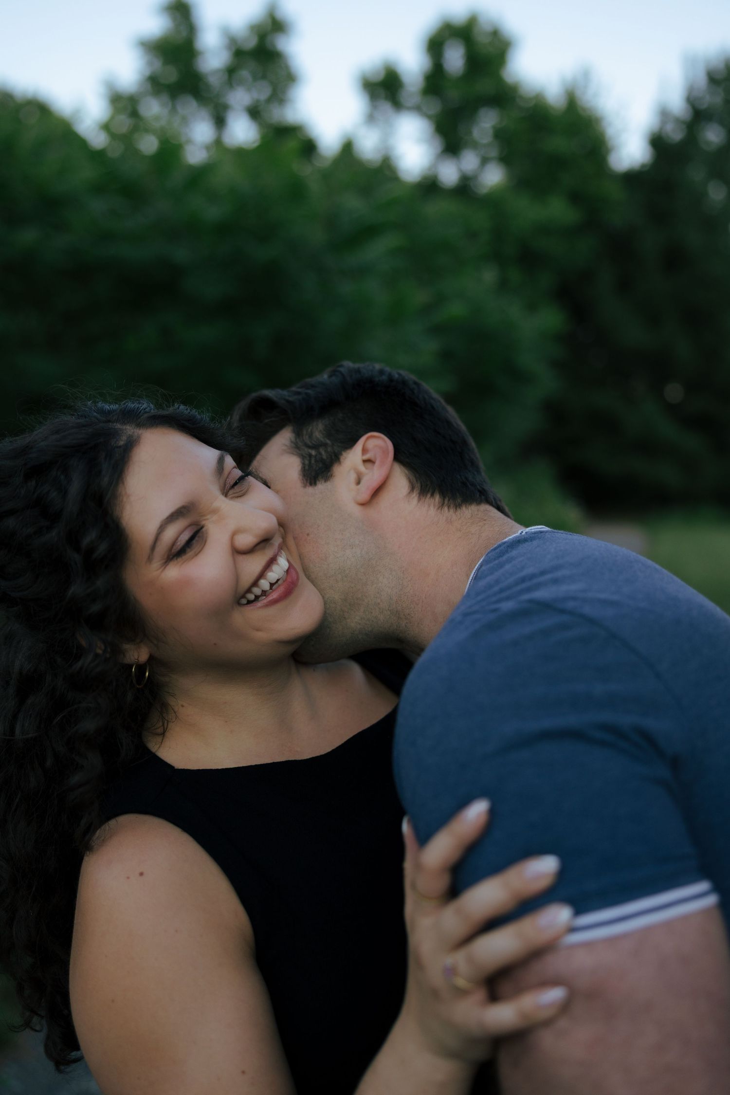 A man kisses a smiling woman on the cheek outdoors, with lush green trees in the background.