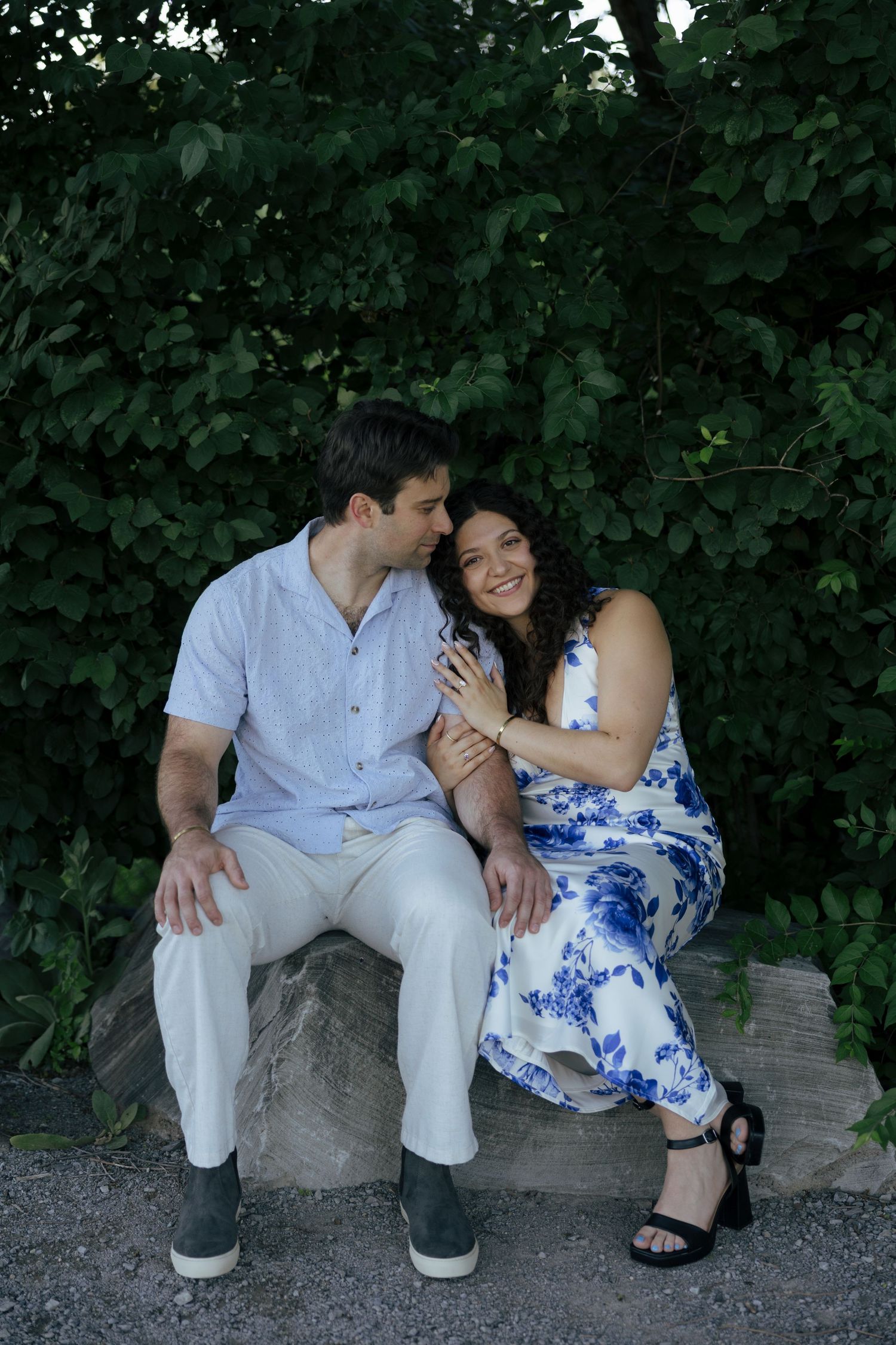 Happy engaged couple sitting on a stone bench embracing and smiling in front of a lush green leafy backdrop.