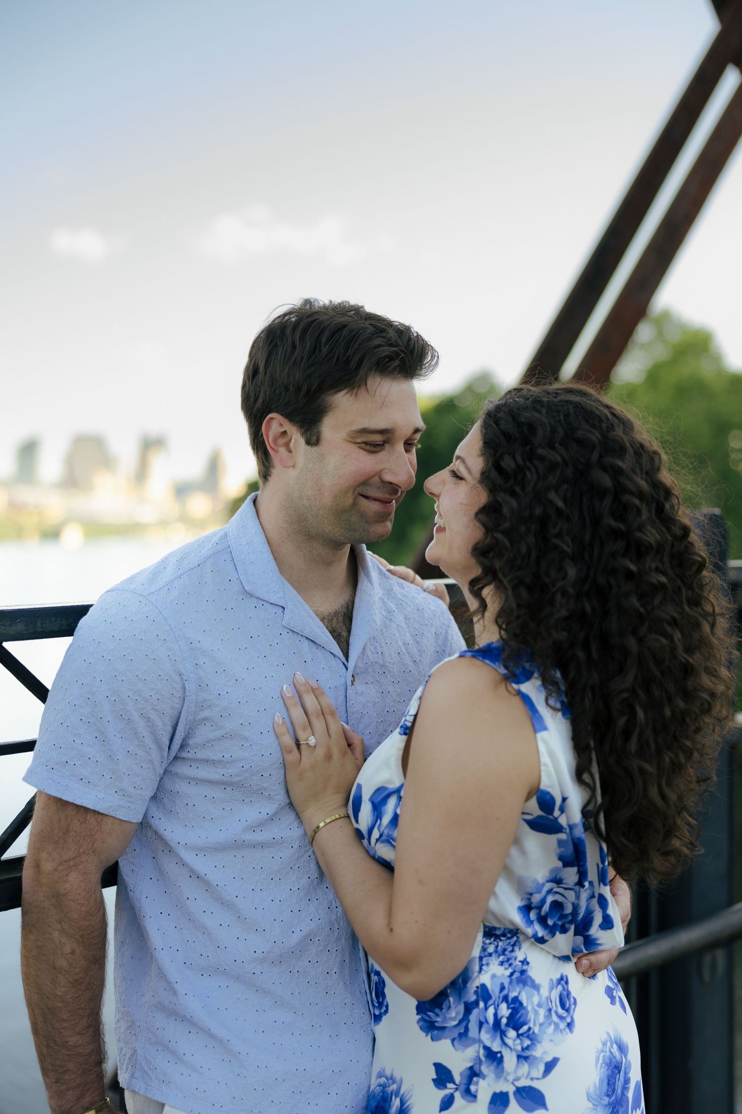 A smiling engaged couple gazes at each other on a bridge, woman wearing a floral blue dress and engagement ring.