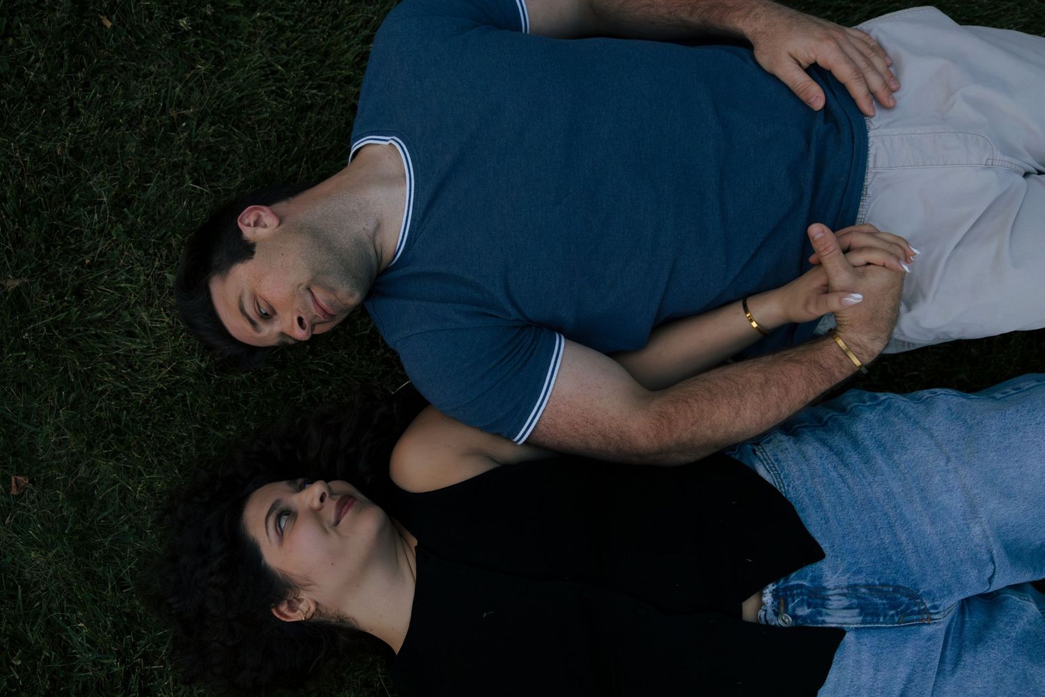 A couple lies on grass holding hands, viewed from above, wearing casual blue and black outfits.