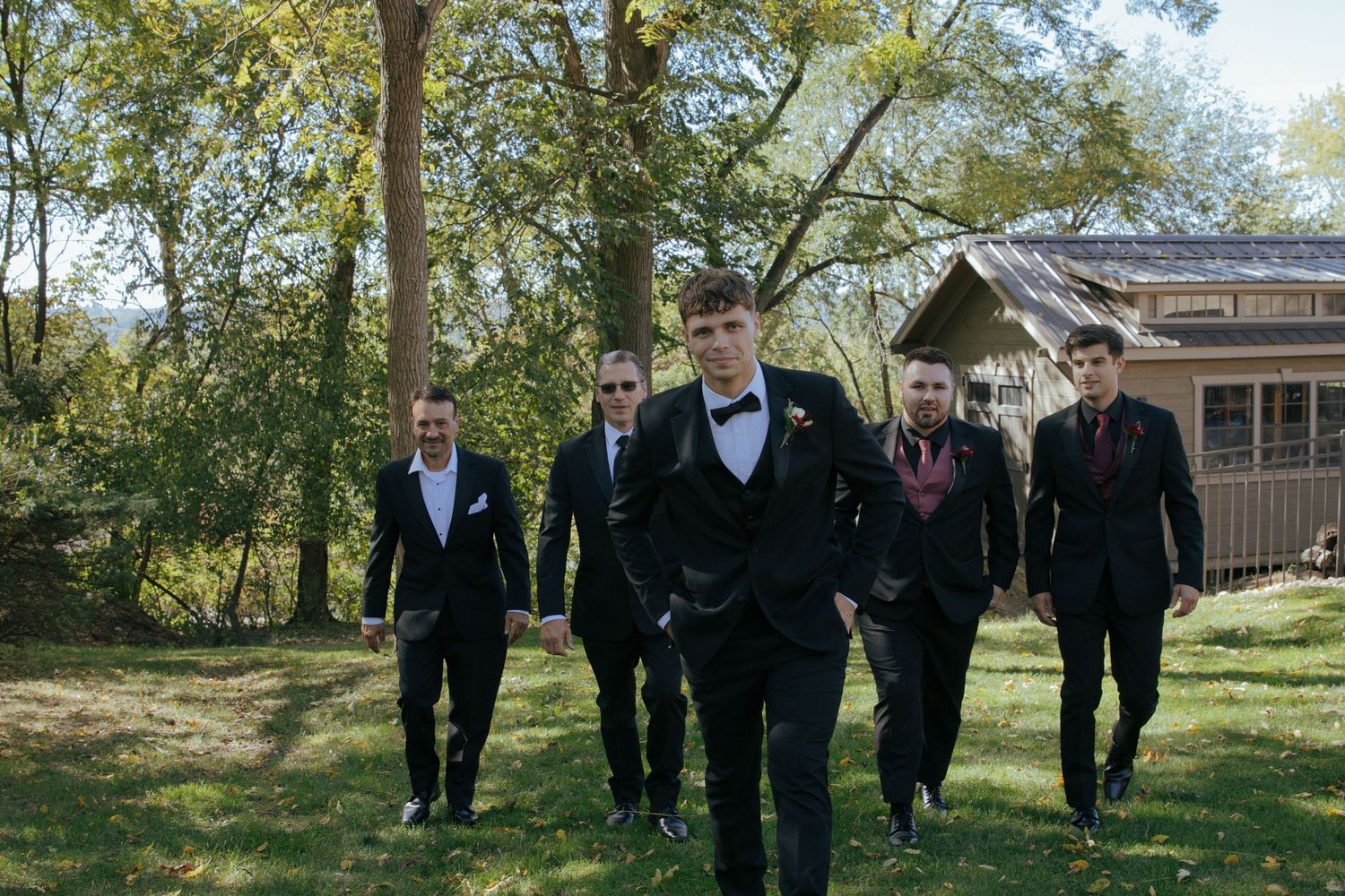 Groom in black tuxedo with bow tie leads groomsmen in dark suits walking outdoors near trees and rustic building.
