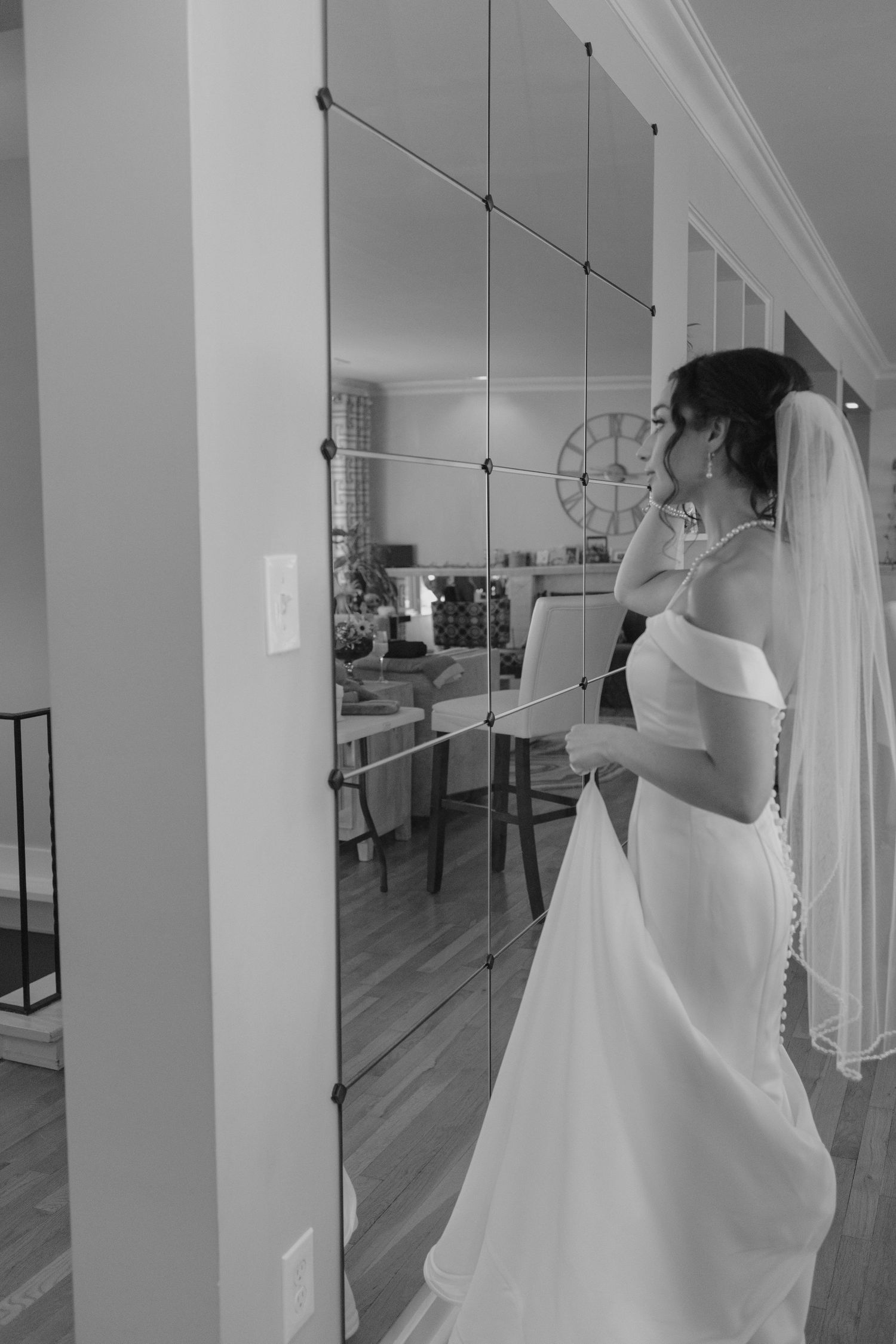 Bride in elegant off-shoulder white gown and veil admires her reflection in a segmented wall mirror indoors.