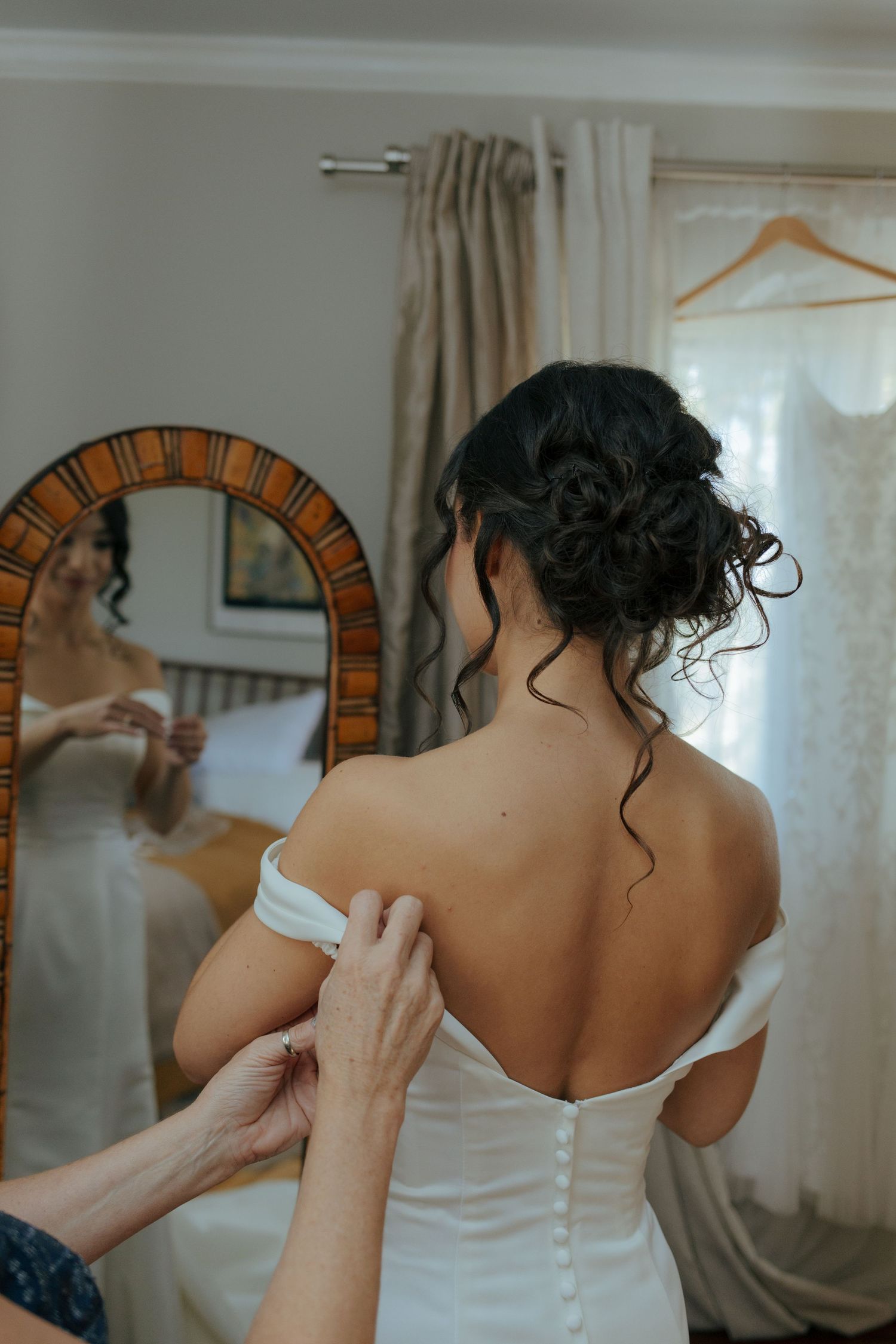 Bride in white button-back wedding gown has dress fastened by assistant before mirror.